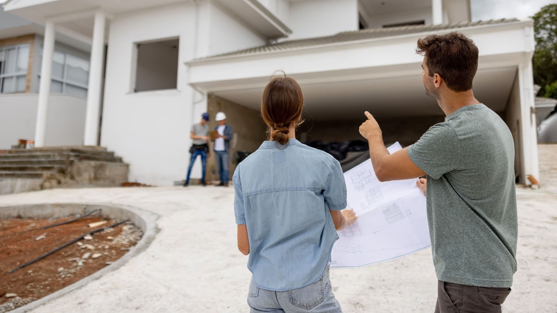 Eine Frau und ein Mann stehen in einer Einfahrt und halten einen Bauplan in den Händen. Der Mann zeigt auf einen noch nicht fertiggestellten Neubau im Hintergrund.