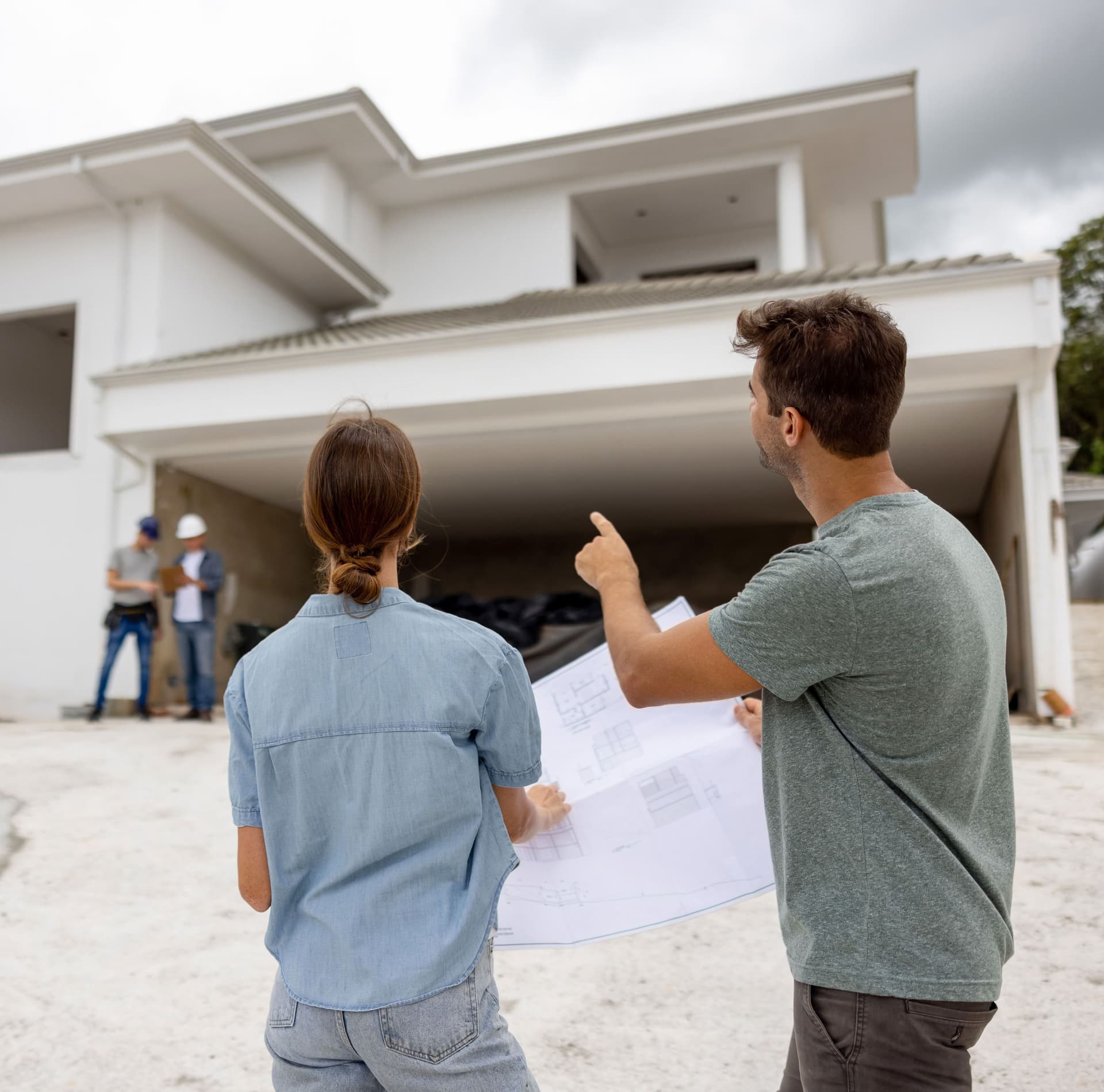 Eine Frau und ein Mann stehen in einer Einfahrt und halten einen Bauplan in den Händen. Der Mann zeigt auf einen noch nicht fertiggestellten Neubau im Hintergrund.