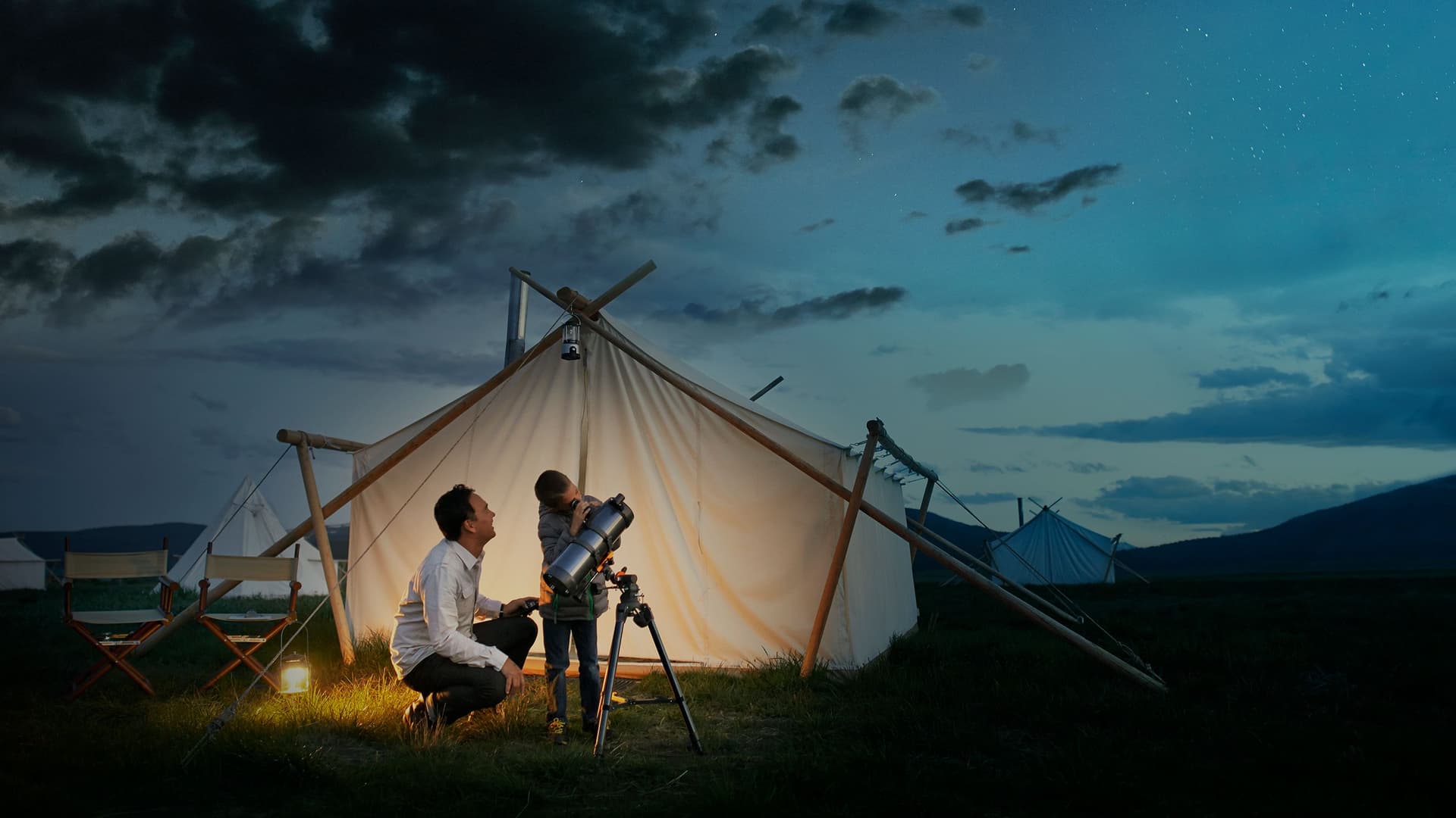 Ein Mann und ein Kind stehen abends vor einem beleuchteten Zelt auf einer Wiese und betrachten gemeinsam den Sternenhimmel durch ein Teleskop. Die Szene wirkt ruhig und idyllisch, umgeben von weiteren Zelten und einem leicht bewölkten Nachthimmel.