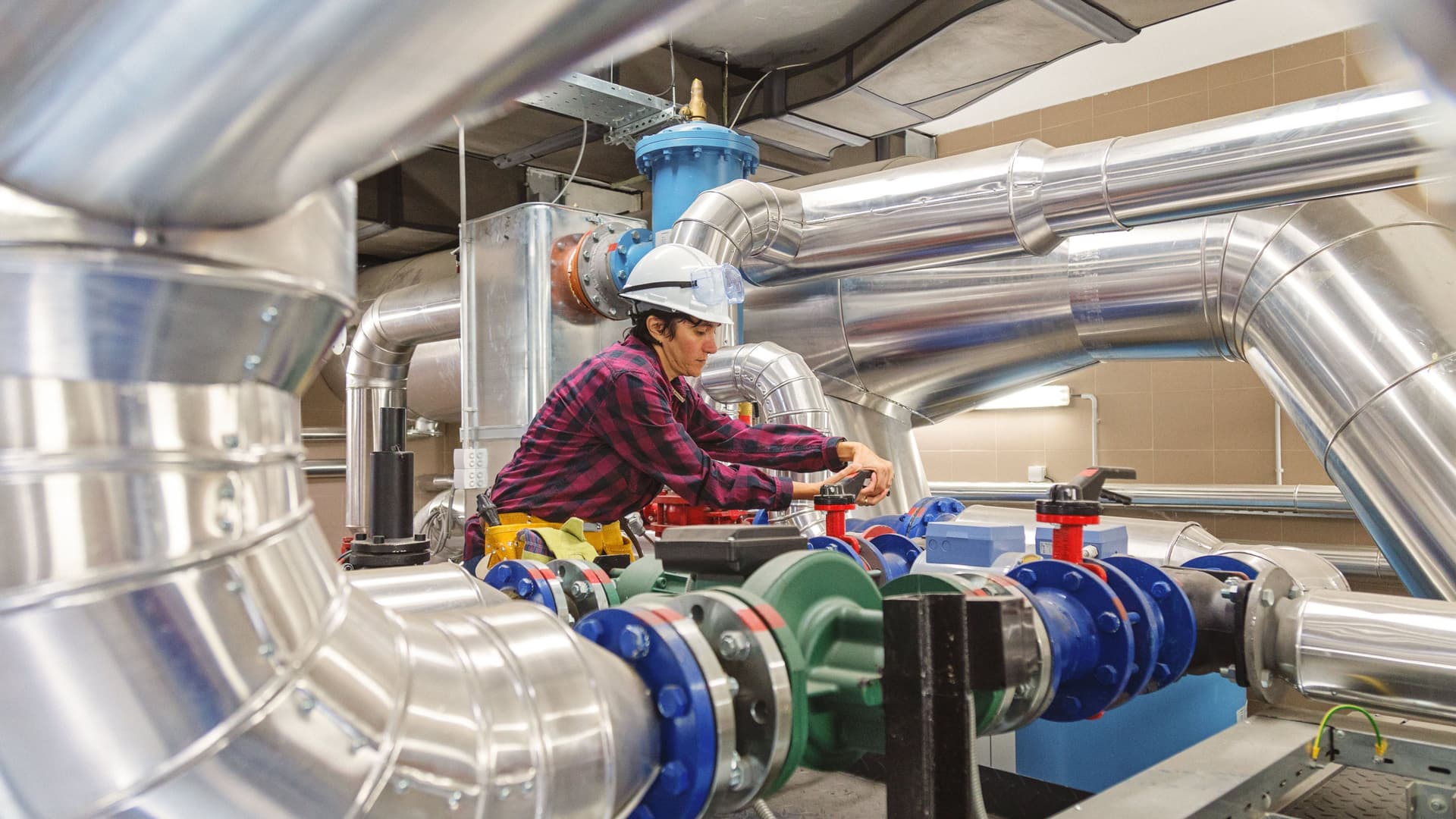 Technikerin mit Schutzhelm und Werkzeuggürtel arbeitet zwischen reflektierenden Metallrohren.