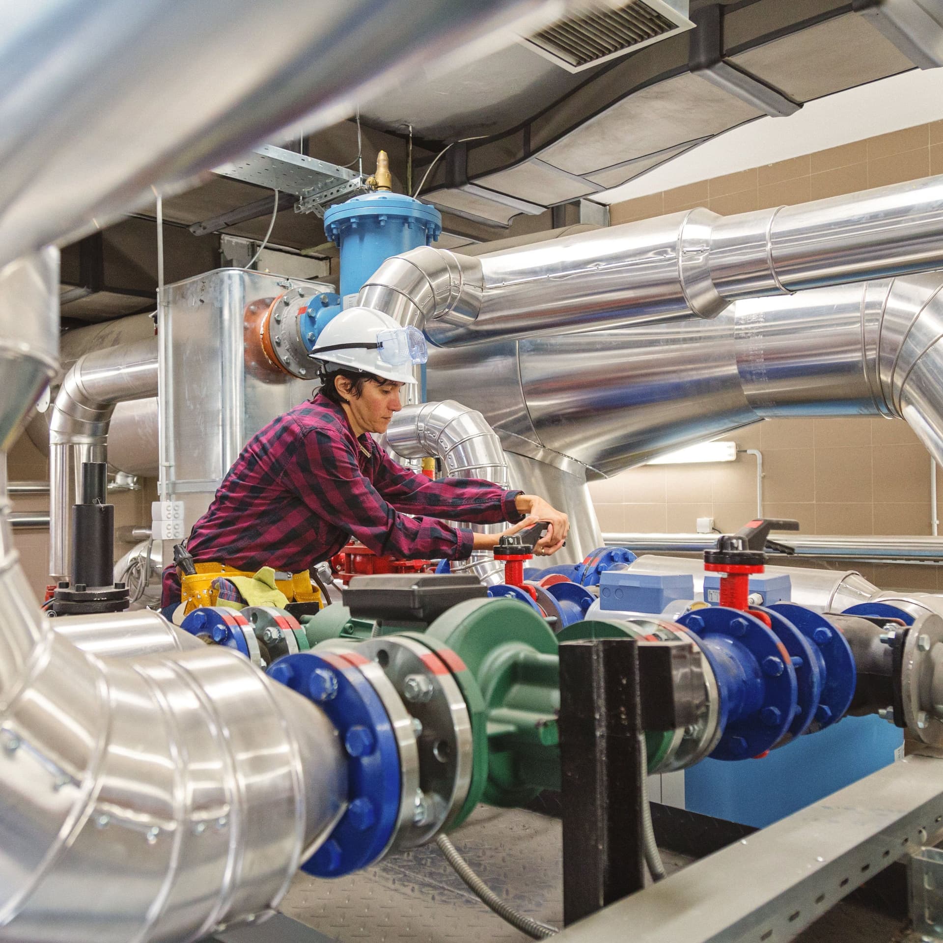 Technikerin mit Schutzhelm und Werkzeuggürtel arbeitet zwischen reflektierenden Metallrohren.