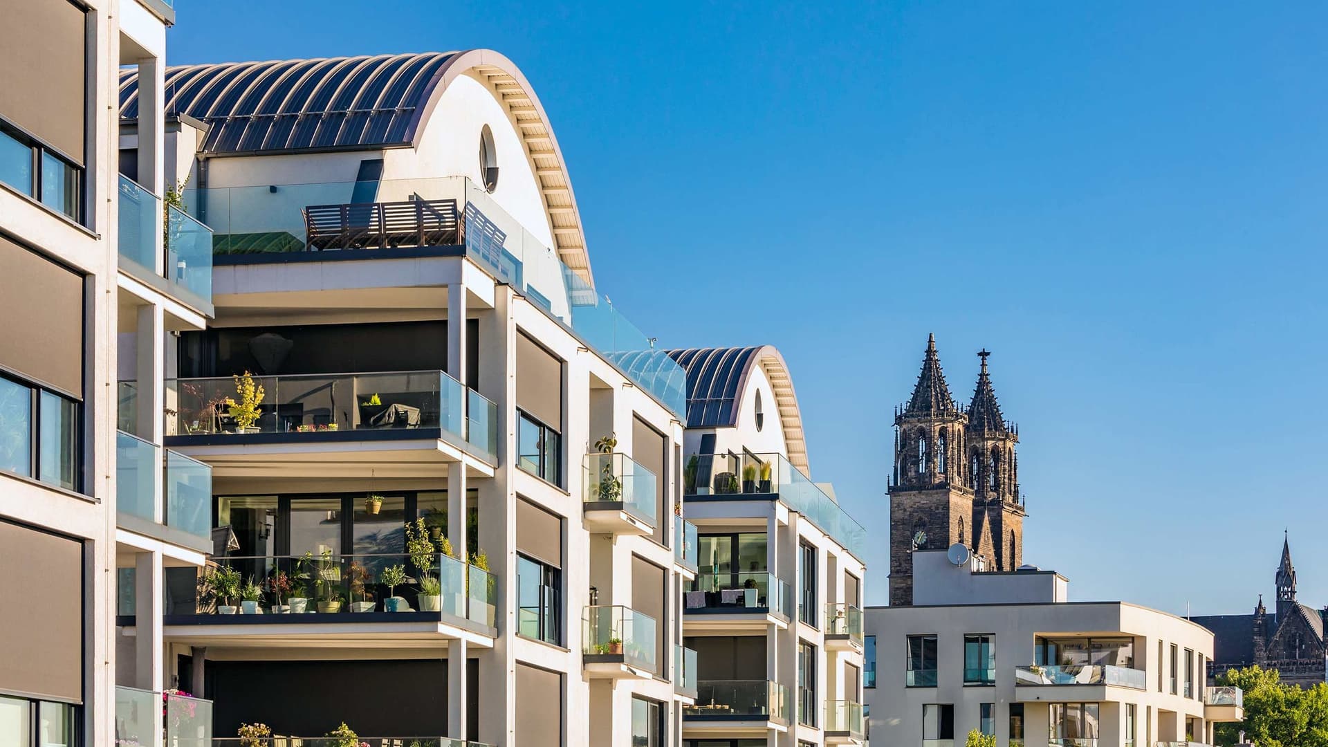 Eine Reihe von modernen Mehrfamilienhäusern in einer Flucht. Hinter dem letzten Gebäude tauchen zwei Türme einer alten Kirche auf. Die Häuser werden von der Sonne angestrahlt und der Himmel ist blau.