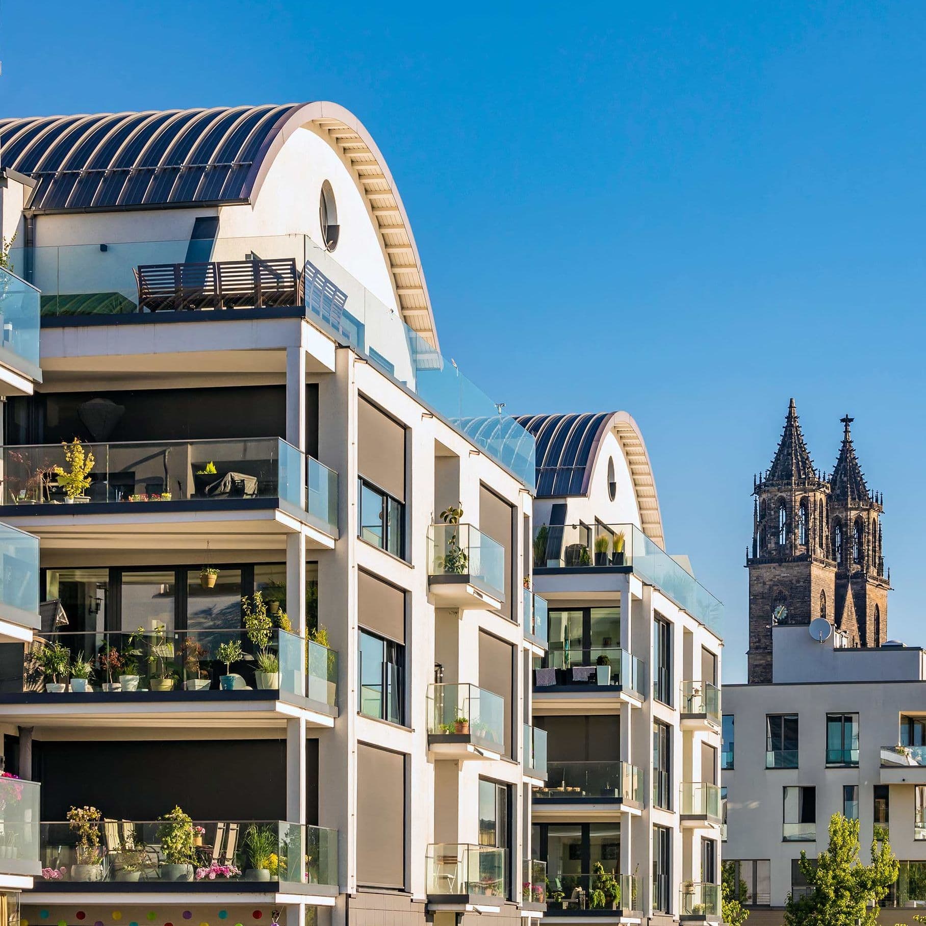 Eine Reihe von modernen Mehrfamilienhäusern in einer Flucht. Hinter dem letzten Gebäude tauchen zwei Türme einer alten Kirche auf. Die Häuser werden von der Sonne angestrahlt und der Himmel ist blau.
