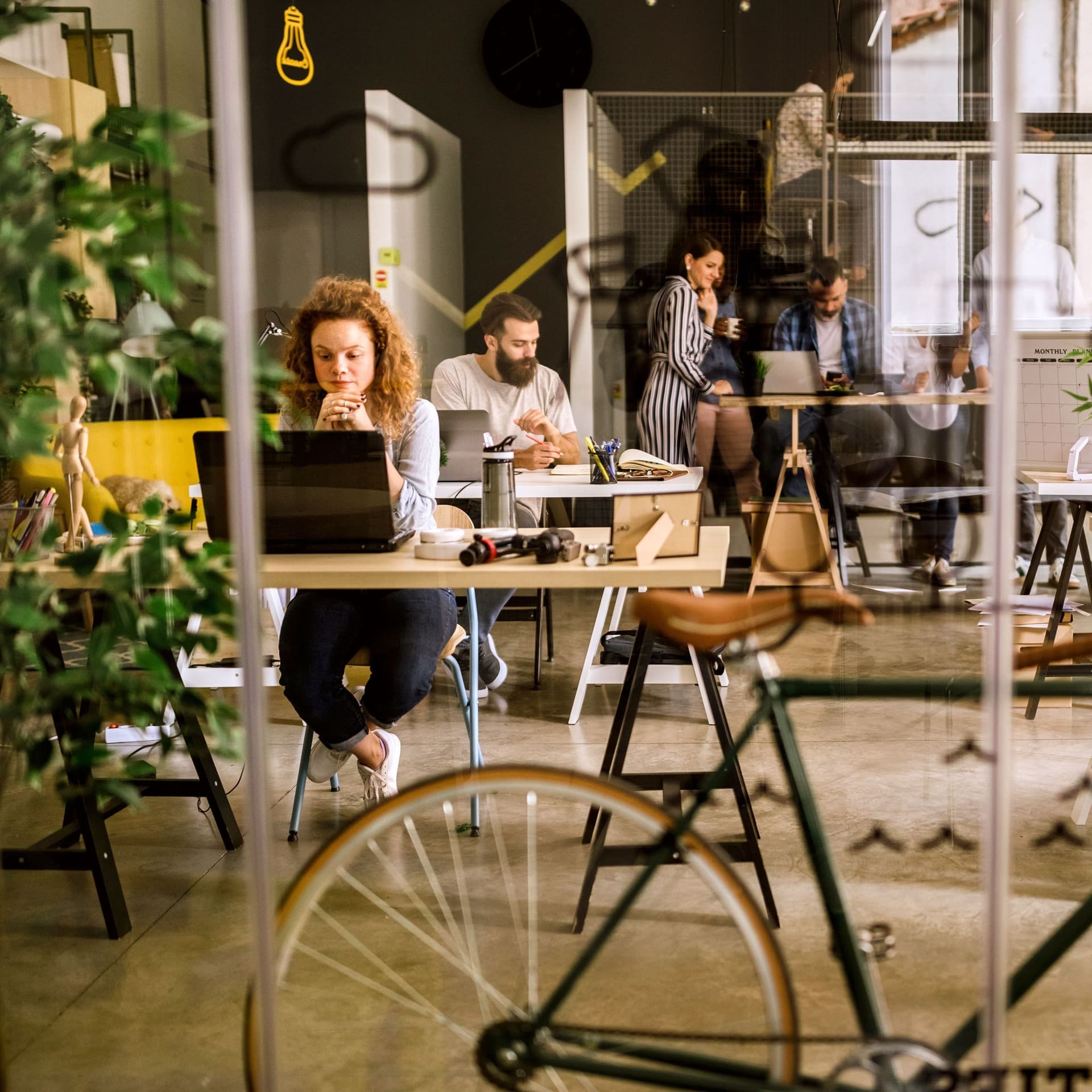 Mehrere Personen in einem hellen begrünten Großraumbüro beim Arbeiten. Im Vordergrund ist ein Fahrrad hinter einer Glasscheibe zu erkennen.