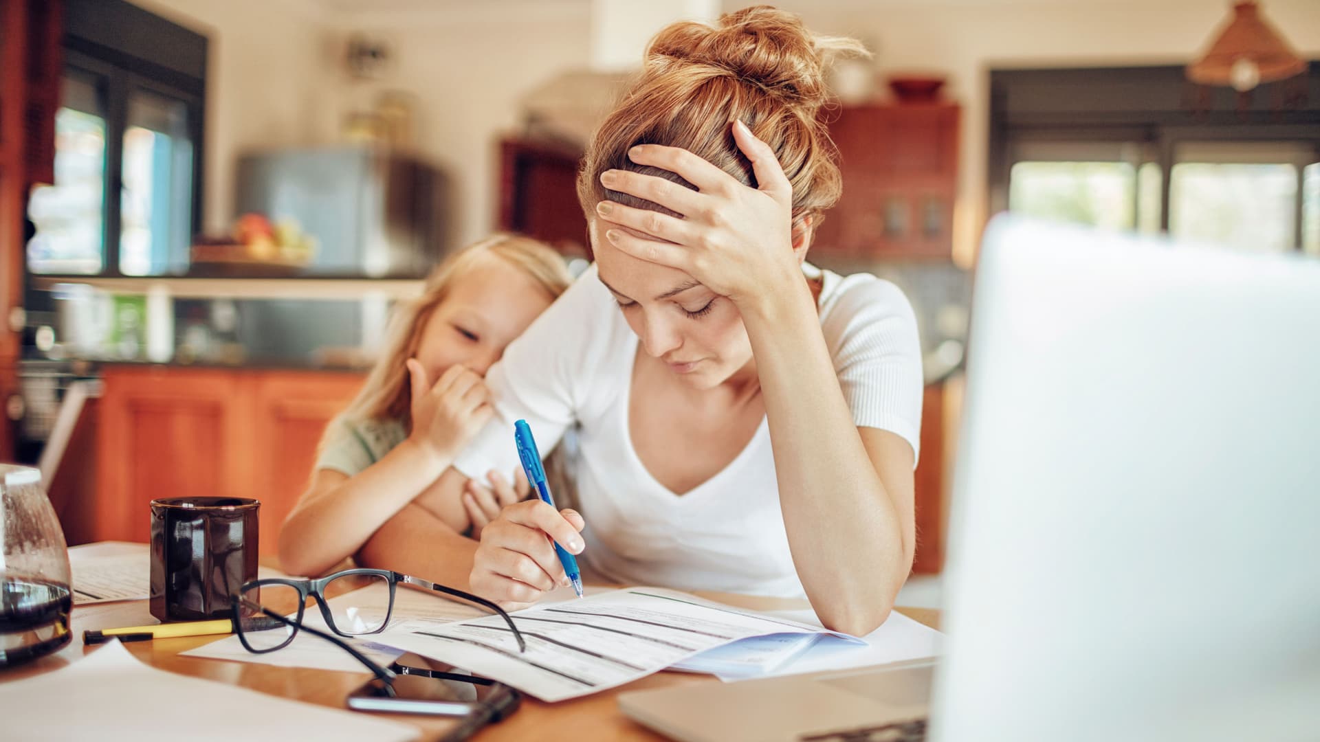 Verzweifelt wirkende junge Mutter mit Kind am Arm sitzt zu Hause vor ihrem Laptop und studiert Unterlagen.