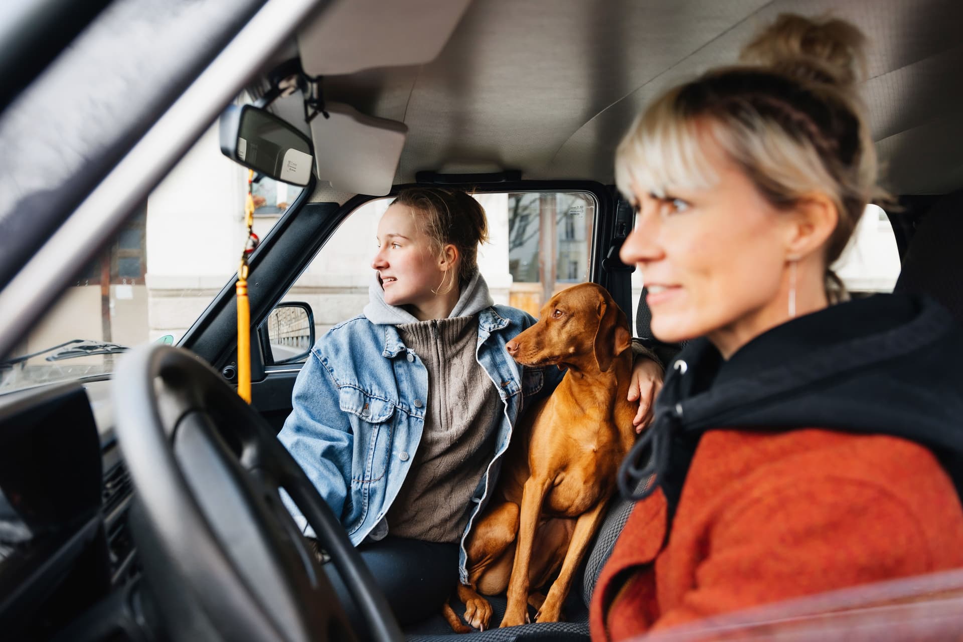 Eine Frau sitzt am Steuer eines Vans während ein Mädchen im Teenageralter und ihr Hund neben ihr sitzen.