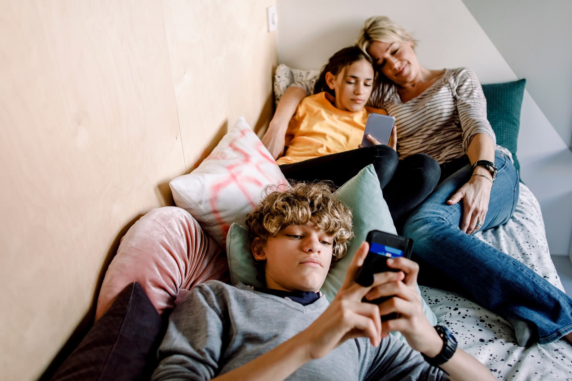 Mutter liegt mit Tochter und Sohn zusammen in einem Bett während diese am Handy sind.