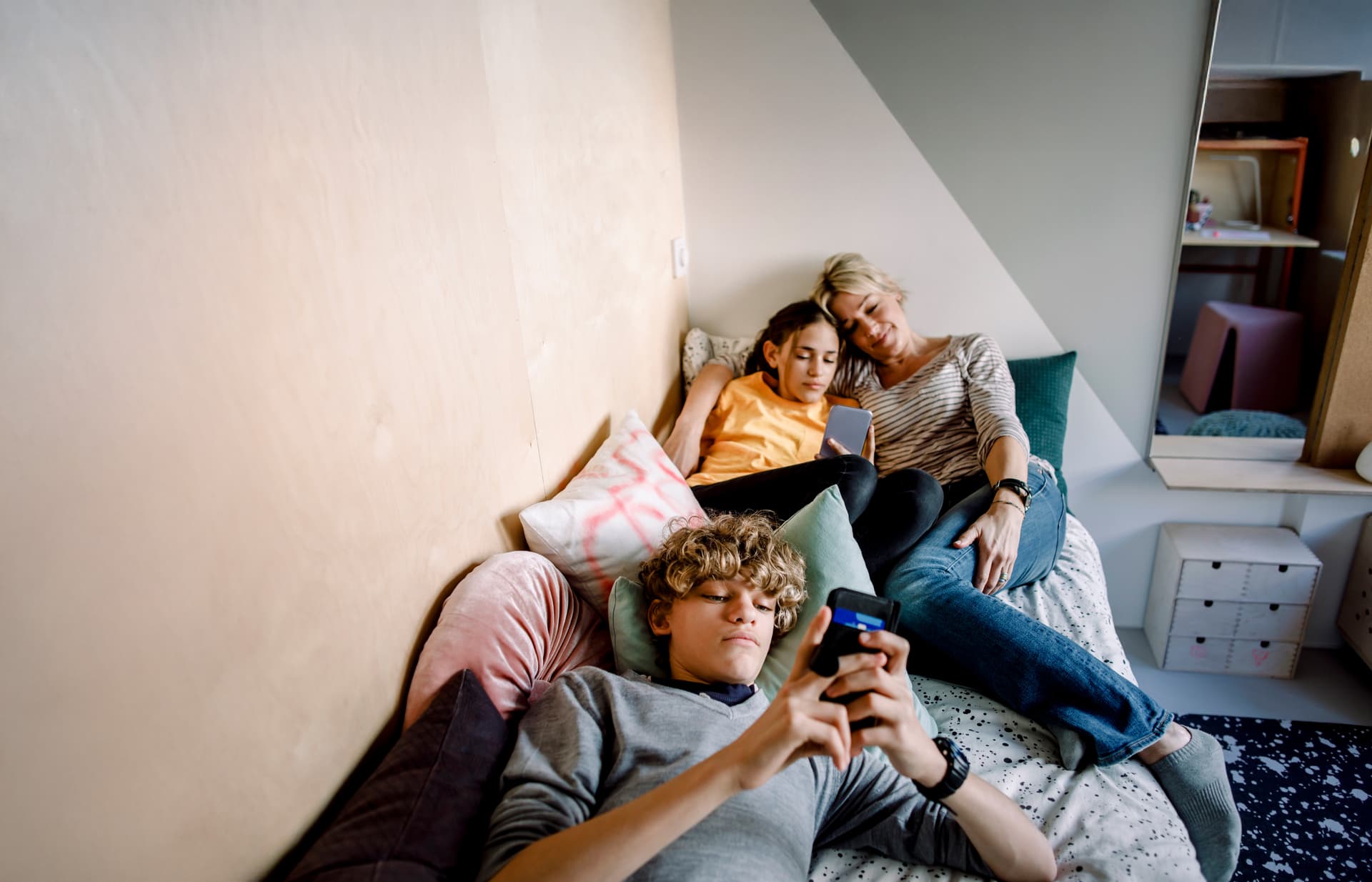 Mutter liegt mit Tochter und Sohn zusammen in einem Bett während diese am Handy sind.