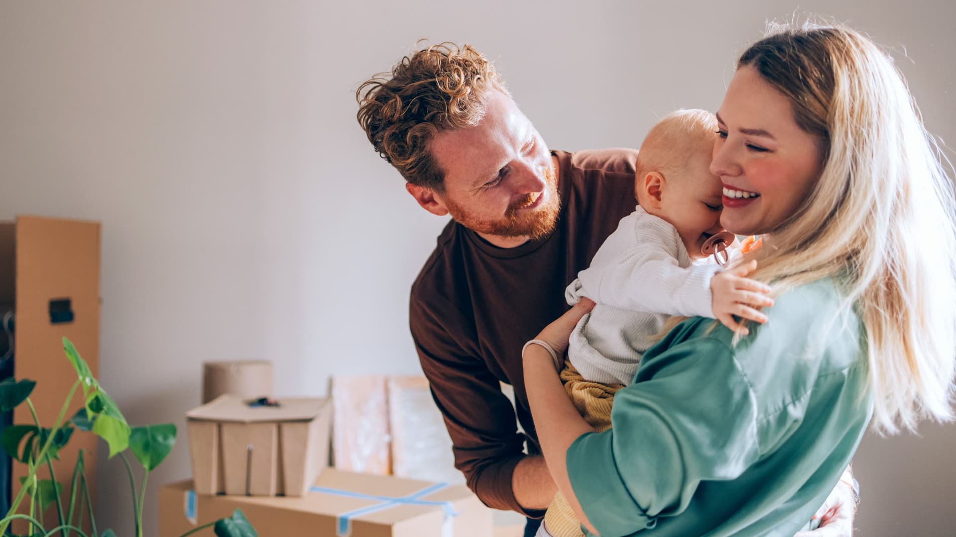 Vater und Mutter mit Baby auf dem Arm stehen lächelnd vor Umzugskartons und einer Zimmerpflanze.