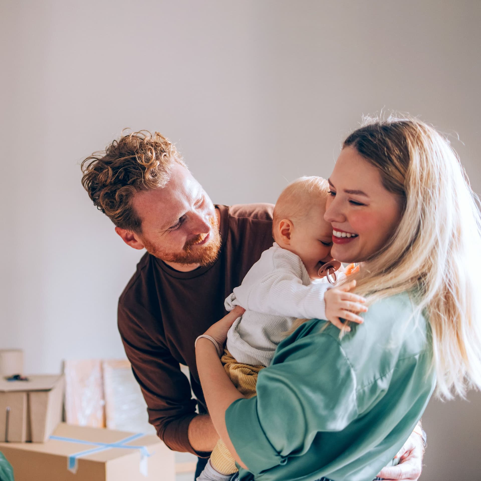 Vater und Mutter mit Baby auf dem Arm stehen lächelnd vor Umzugskartons und einer Zimmerpflanze.