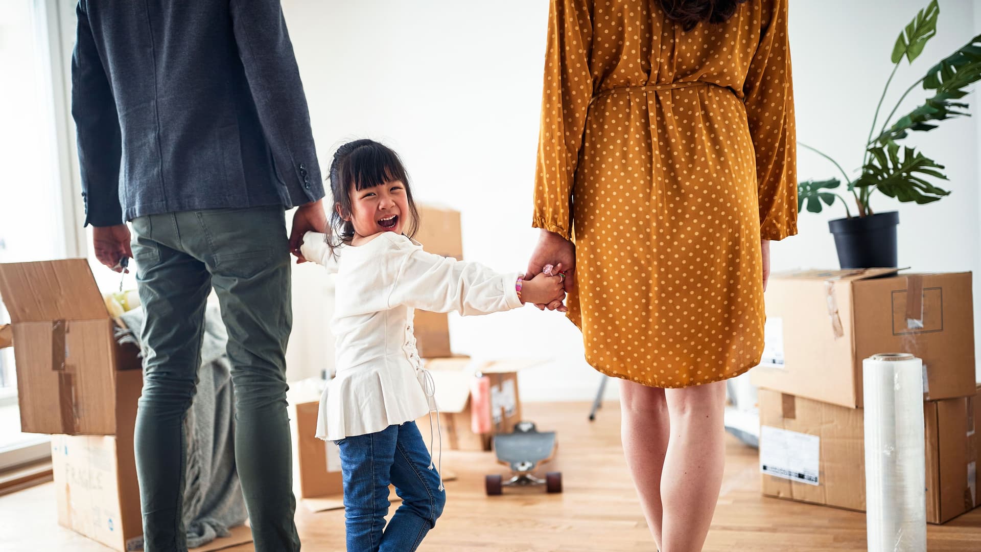 Lachende Kind an den Händen einer Frau und eines Mannes, die mit dem Rücken zur Kamera stehen. Sie stehen in einer Wohnung mit Umzugskisten.