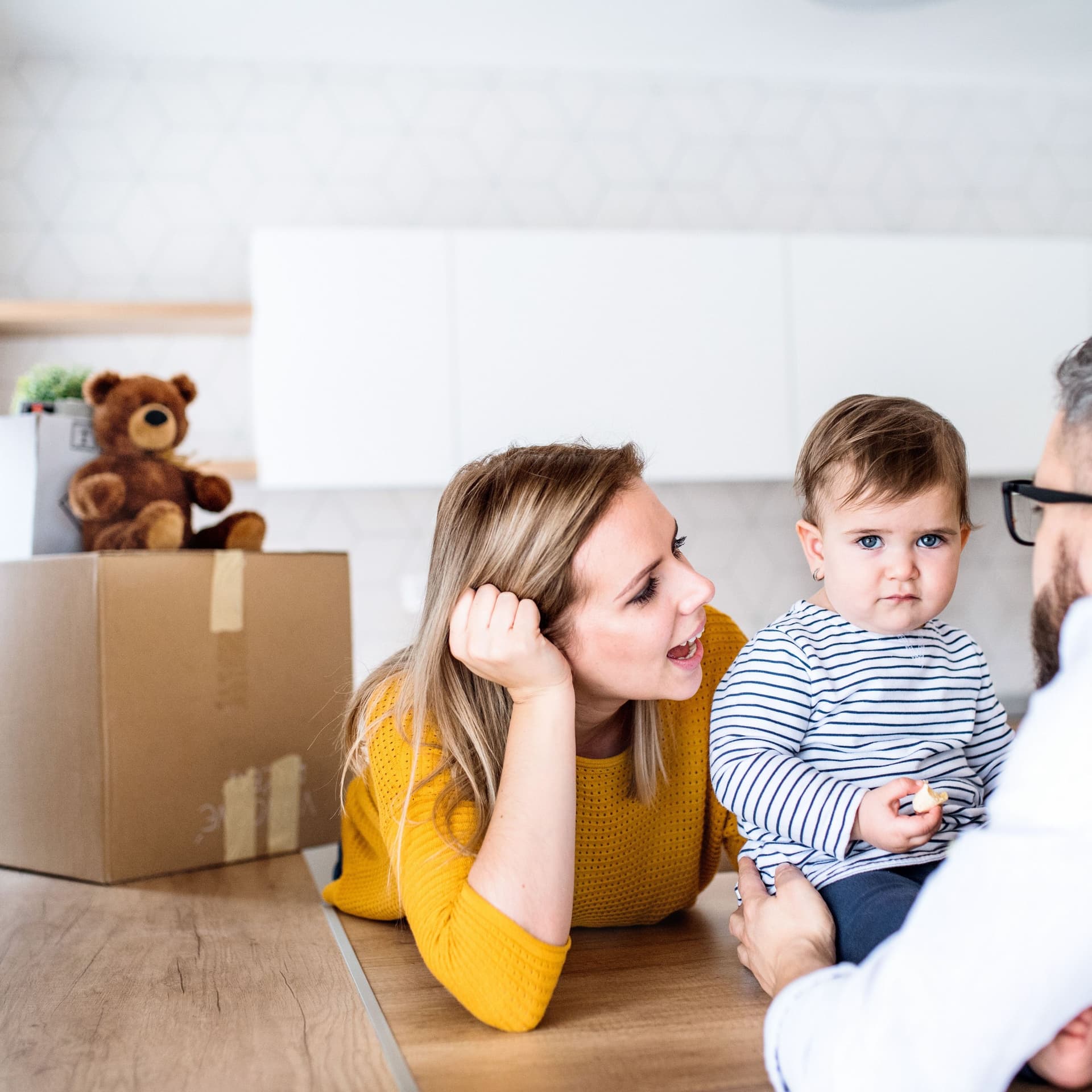 Ein Porträt einer jungen Familie mit einem Kleinkind, dass auf einem Holztisch sitzt. Es wird gehalten von dem Vater, der mit dem Rücken zur Kamera steht.