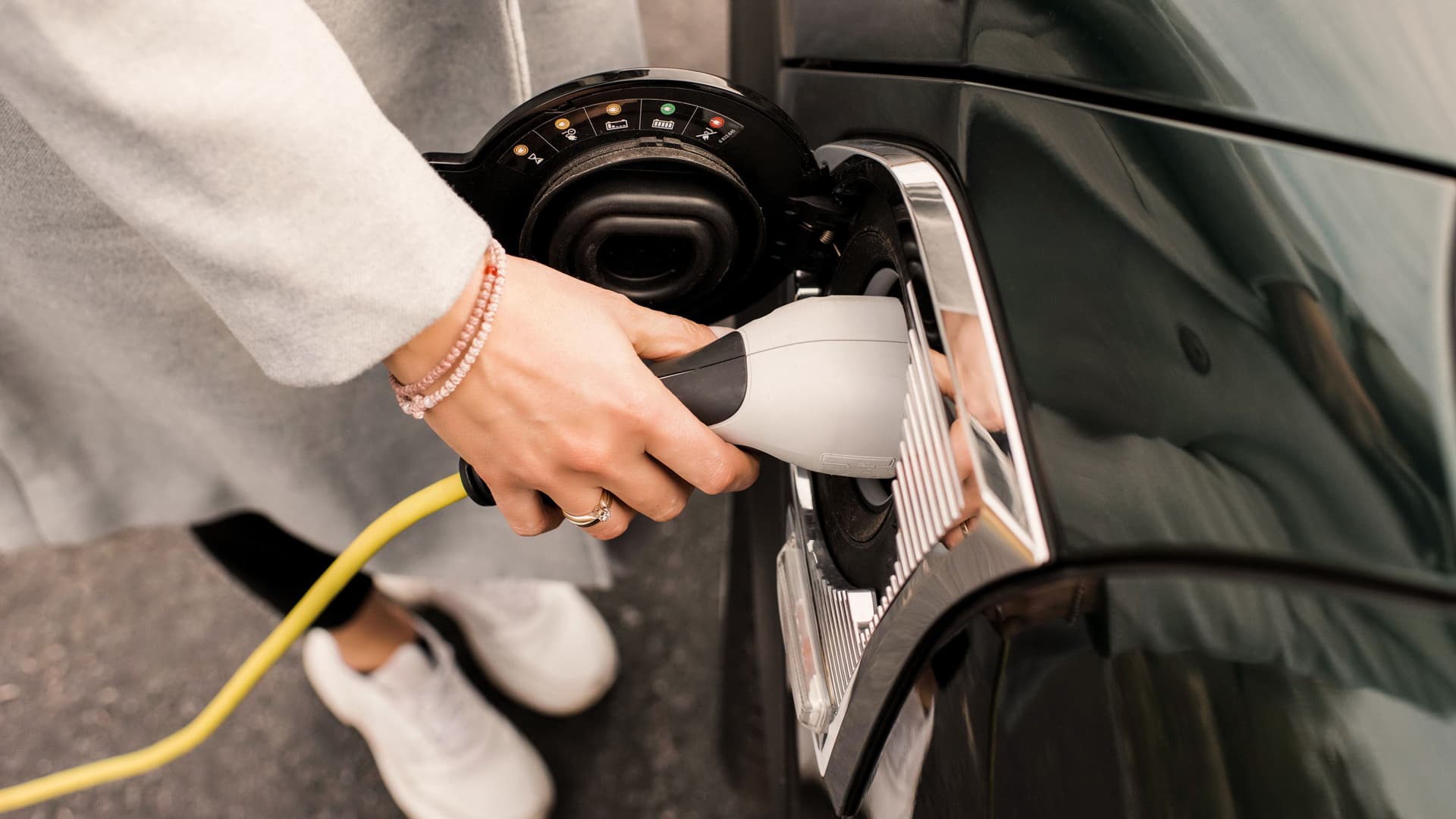 Nahaufnahme einer Hand einer Frau mit weißen Turnschuhen, die ein Ladekabel an ihr Elektroauto anschließt.