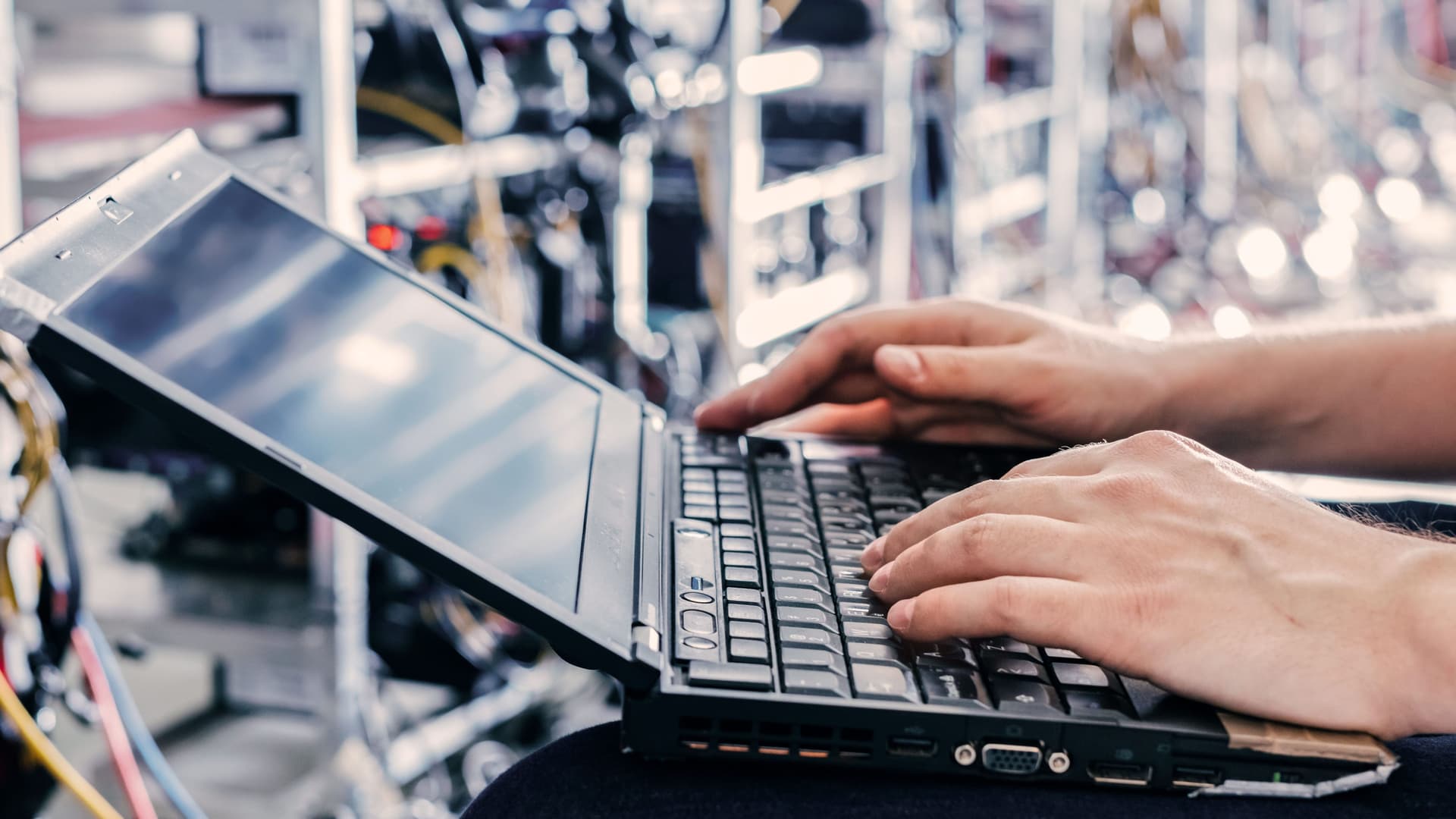 Nahaufnahme einer Person mit Laptop auf den Knien, die in einem Serverraum sitzt.