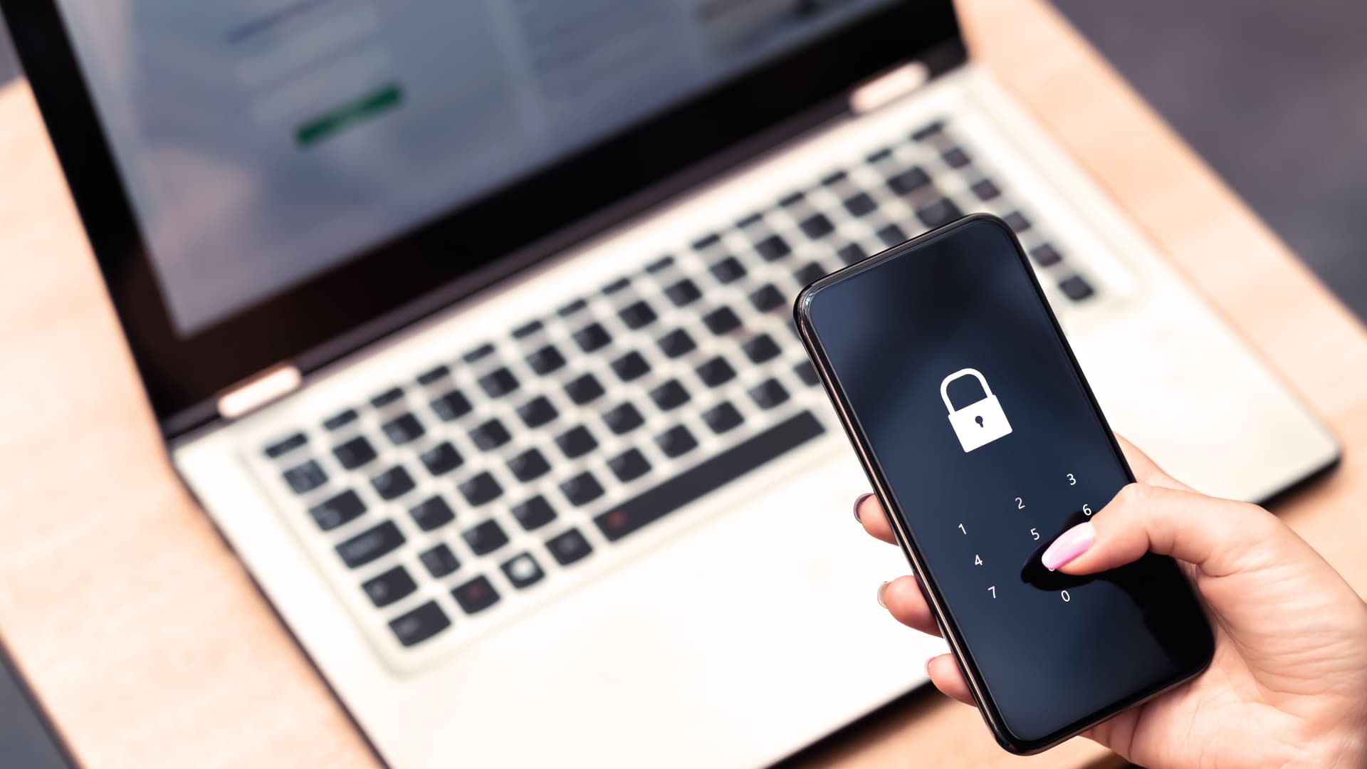Nahaufname einer Hand mit Smartphone vor Laptop. Auf dem Display ist ein Vorhängeschloss abgebildet.