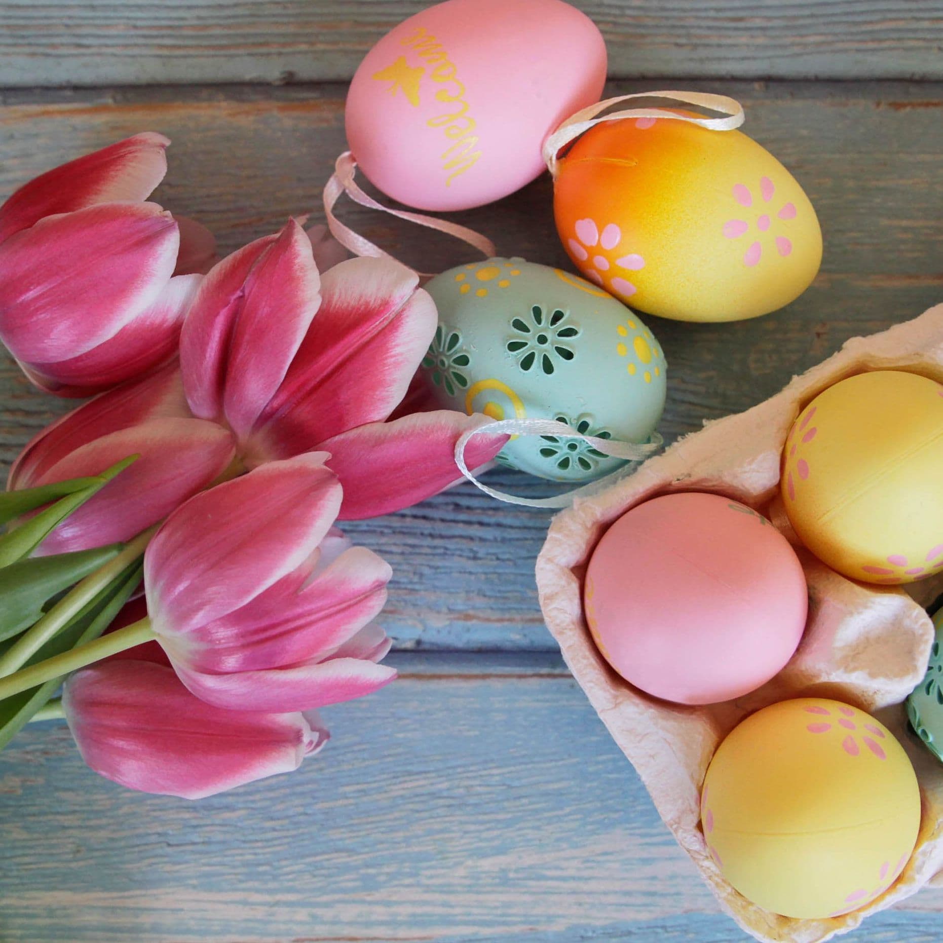 Neues im April: Ein Eierkarton gefüllt mit bunten Eiern liegt auf einem Tisch, daneben ein Strauß rosa Tulpen. 