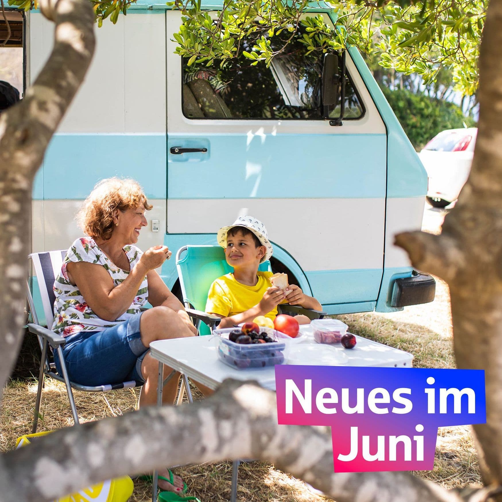 Eine Frau und ein Junge frühstücken vor einem Campingbus. Die Sonne scheint. Darauf steht in einem bunten Feld "Neues im Juni"