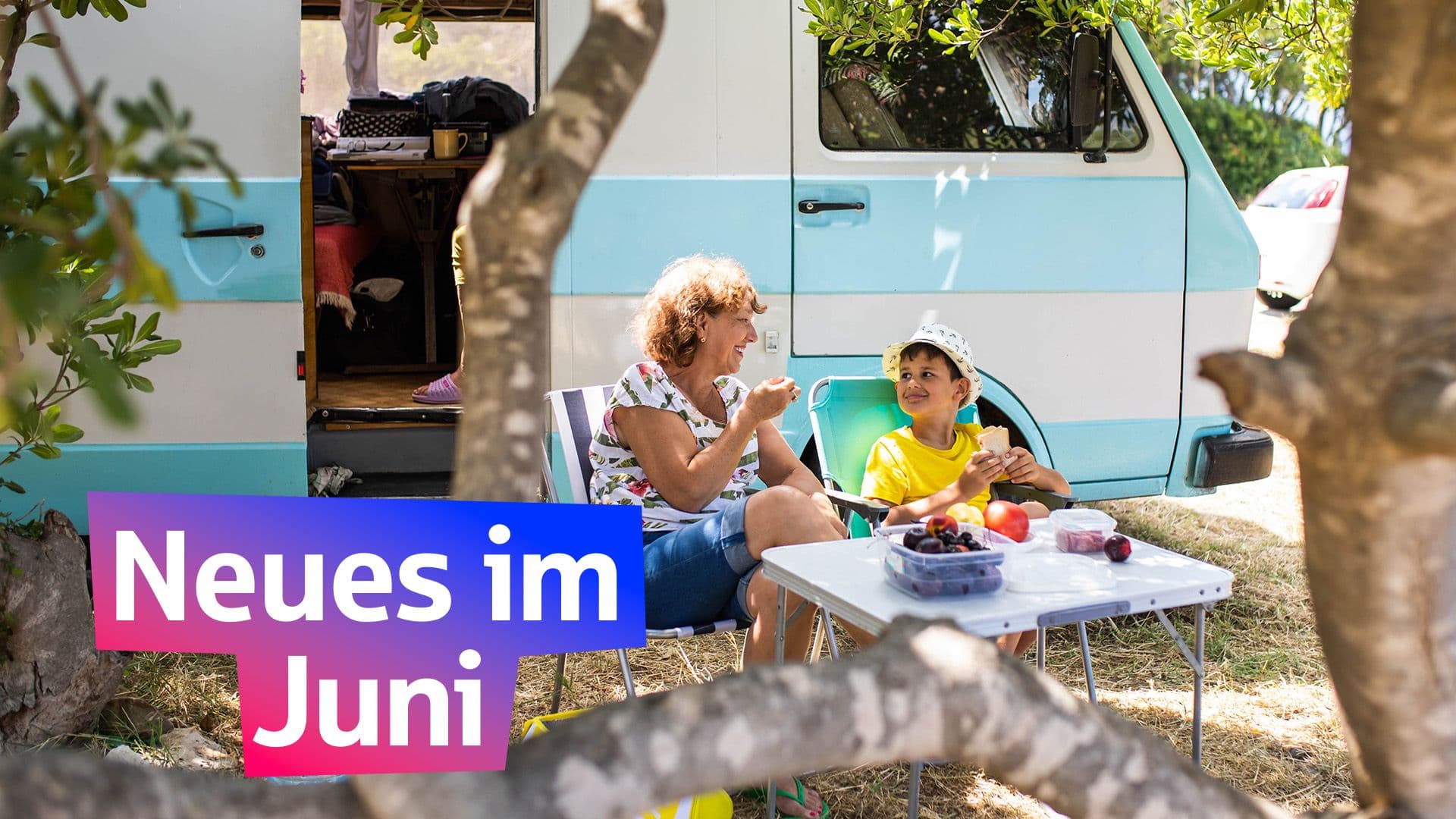 Eine Frau und ein Junge frühstücken vor einem Campingbus. Die Sonne scheint. Darauf steht in einem bunten Feld "Neues im Juni"