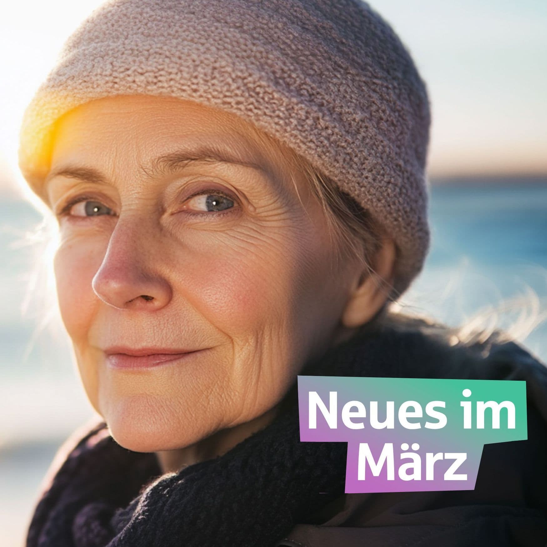 Eine ältere Frau steht vor dem Meer und schaut in die Kamera. Sie hat eine Mütze auf.