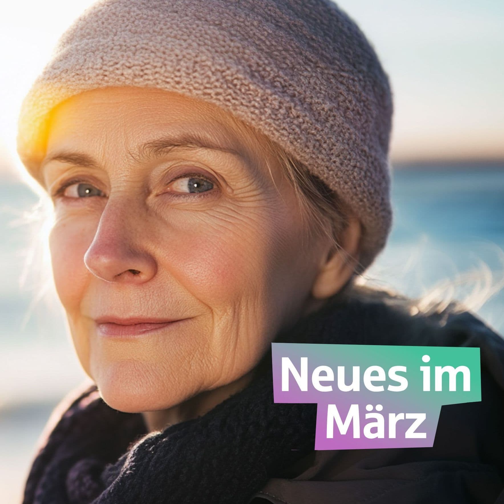 Eine ältere Frau steht vor dem Meer und schaut in die Kamera. Sie hat eine Mütze auf.