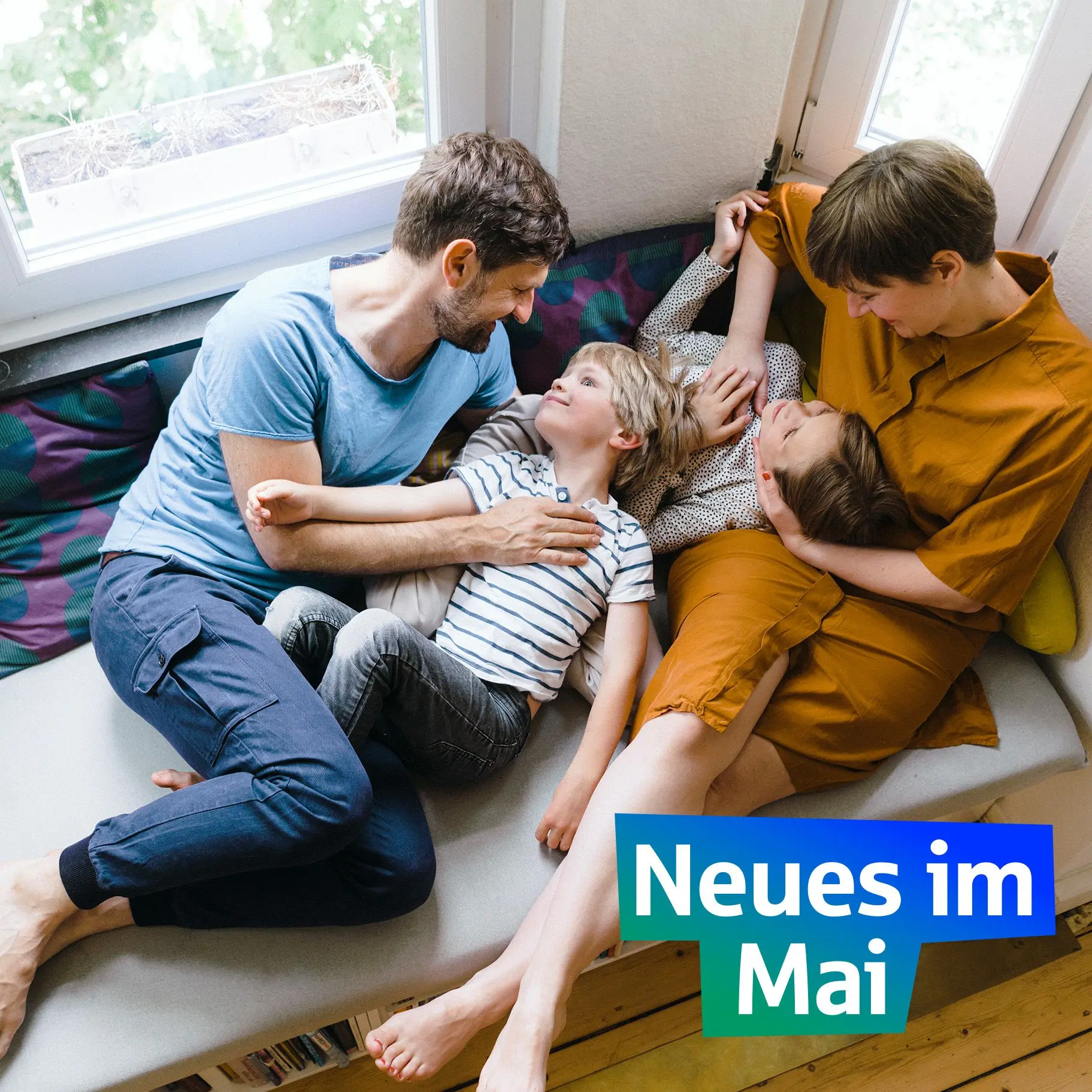 Eine Familie liegt kuschelnd auf ihrer Couch am Fenster.