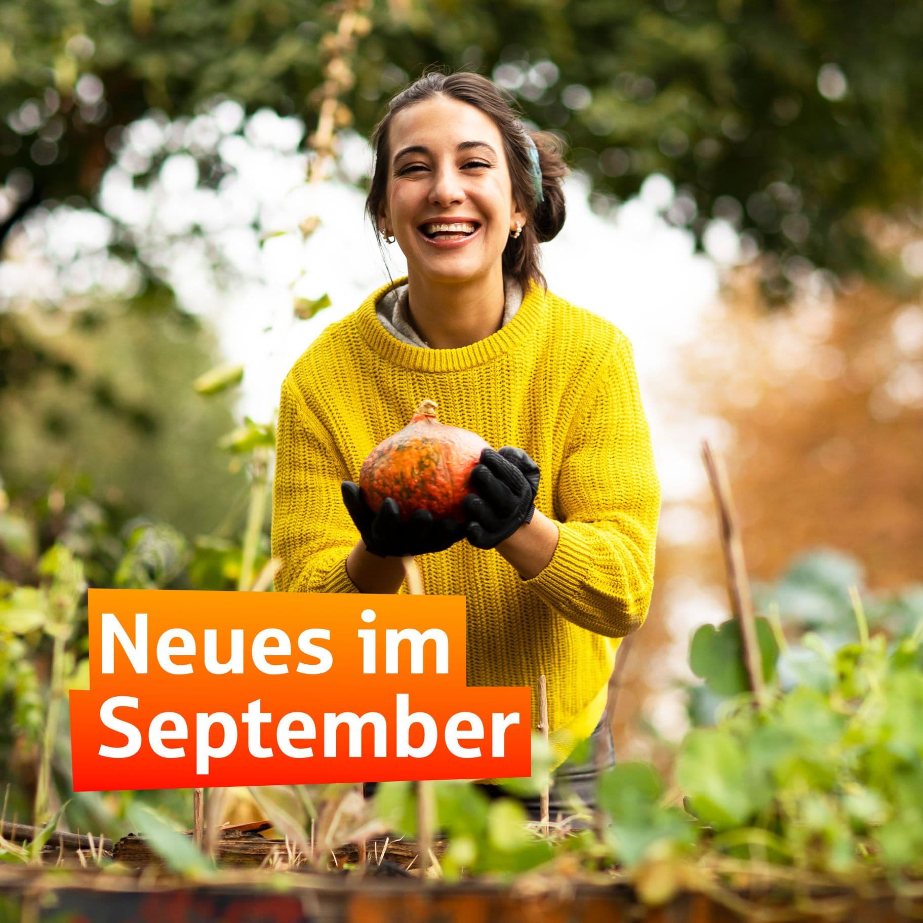 Eine junge Frau mit gelbem Pullover und Kopfhörern in ihrem Garten, lächelt fröhlich in die Kamera und hält einen Kürbis in beiden Händen.