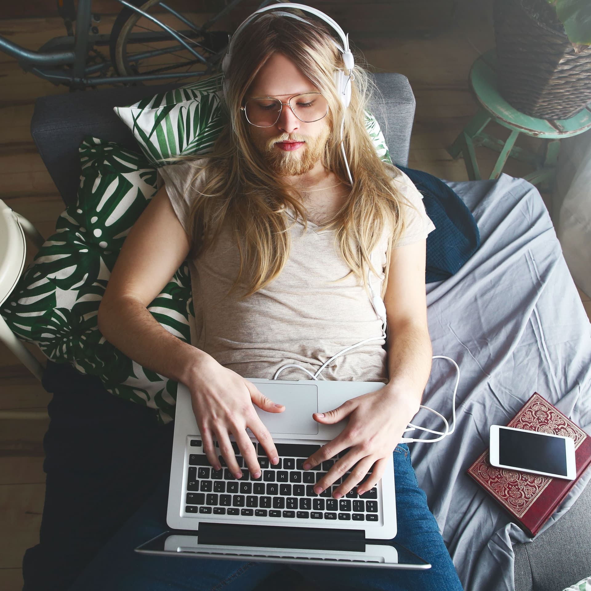 Ein Mann mit langen Haaren und Bart liegt auf einem Sofa. Er trägt Kopfhörer und hat einen Laptop auf seinem Bauch.