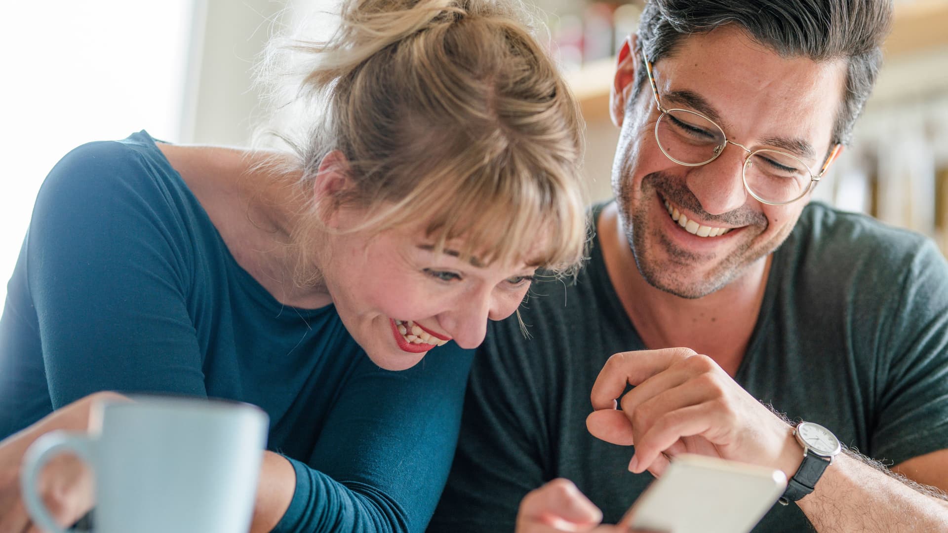 Ein Paar mittleren Alters sitzt zusammen und schaut auf ein Smartphone. Sie lachen ausgelassen.