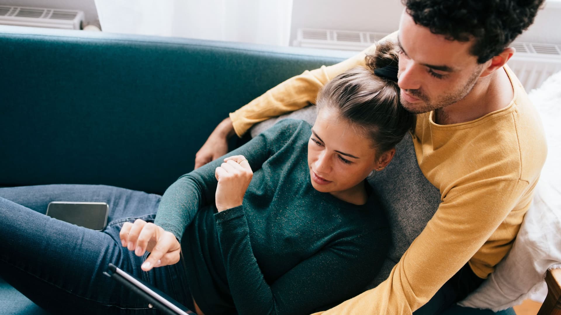 Ein Mann und eine Frau benutzen ein Tablet, während sie sich zu Hause auf dem Sofa liegen. Der Mann umarmt die Frau mit seinem Armen von hinten.