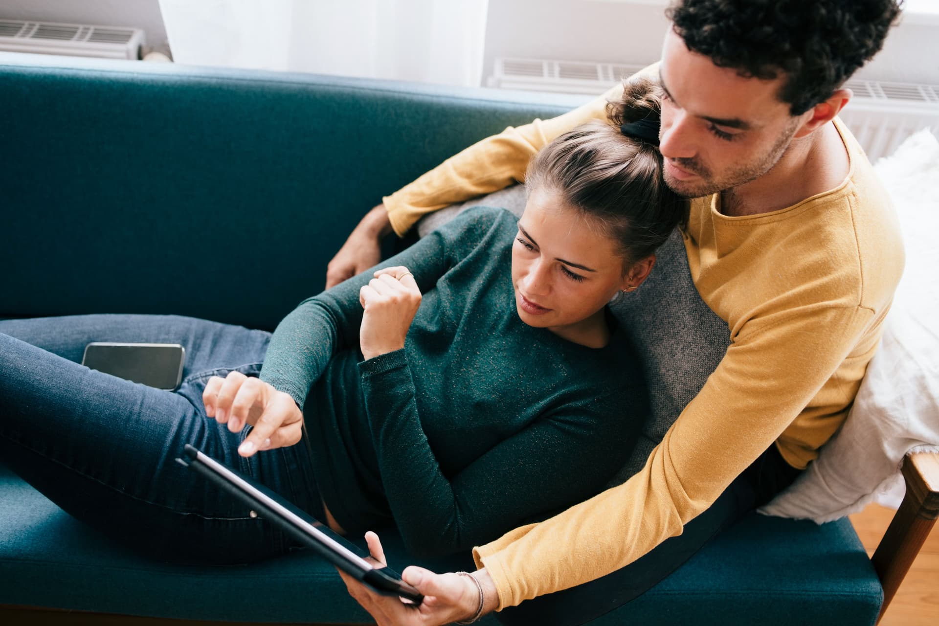 Ein Mann und eine Frau benutzen ein Tablet, während sie sich zu Hause auf dem Sofa liegen. Der Mann umarmt die Frau mit seinem Armen von hinten.