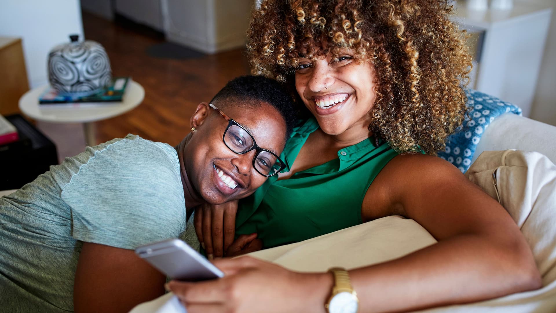 zwei lächelnde Frauen, die zusammen auf einem Sofa sitzen und kuscheln. Die eine Frau hält ein Smartphone in der Hand.