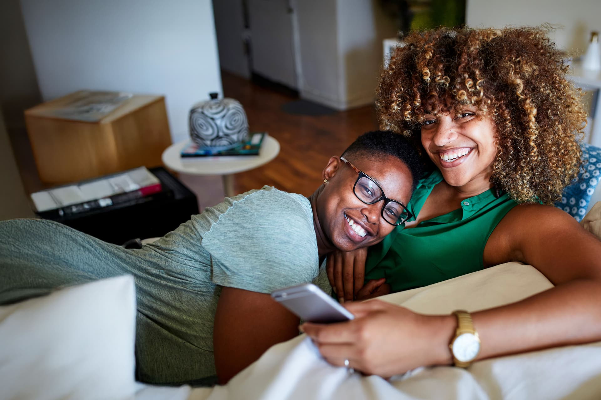 zwei lächelnde Frauen, die zusammen auf einem Sofa sitzen und kuscheln. Die eine Frau hält ein Smartphone in der Hand.