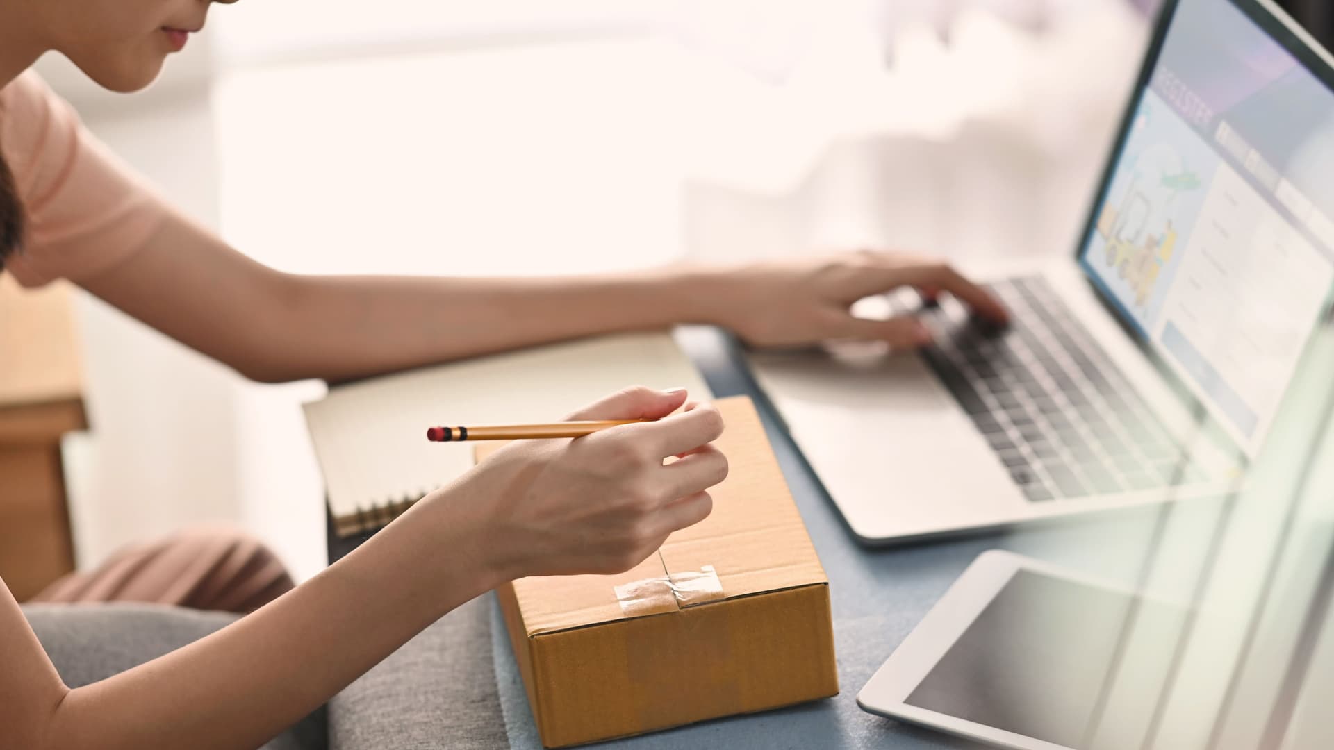 Ausschnitt junge Frau die mit der einen Hand ein Paket beschriftet und mit der anderen den Laptop bedient.