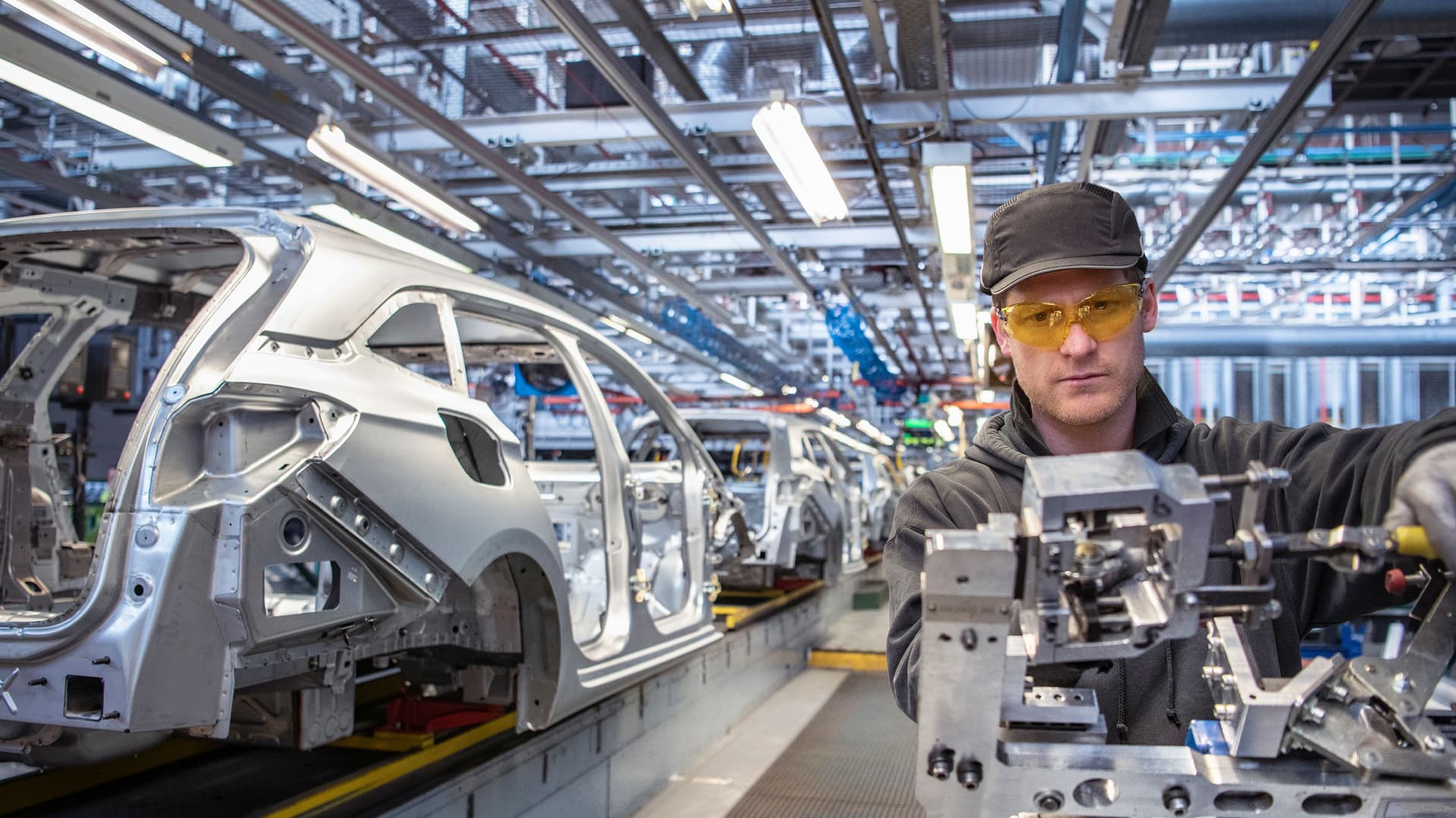  Arbeiter in Schutzkleidung bedient ein Montagewerkzeug an einer Fahrzeugkarosserielinie in einer hell beleuchteten Automobilfabrik, im Hintergrund stehen mehrere Rohkarossen auf dem Fließband