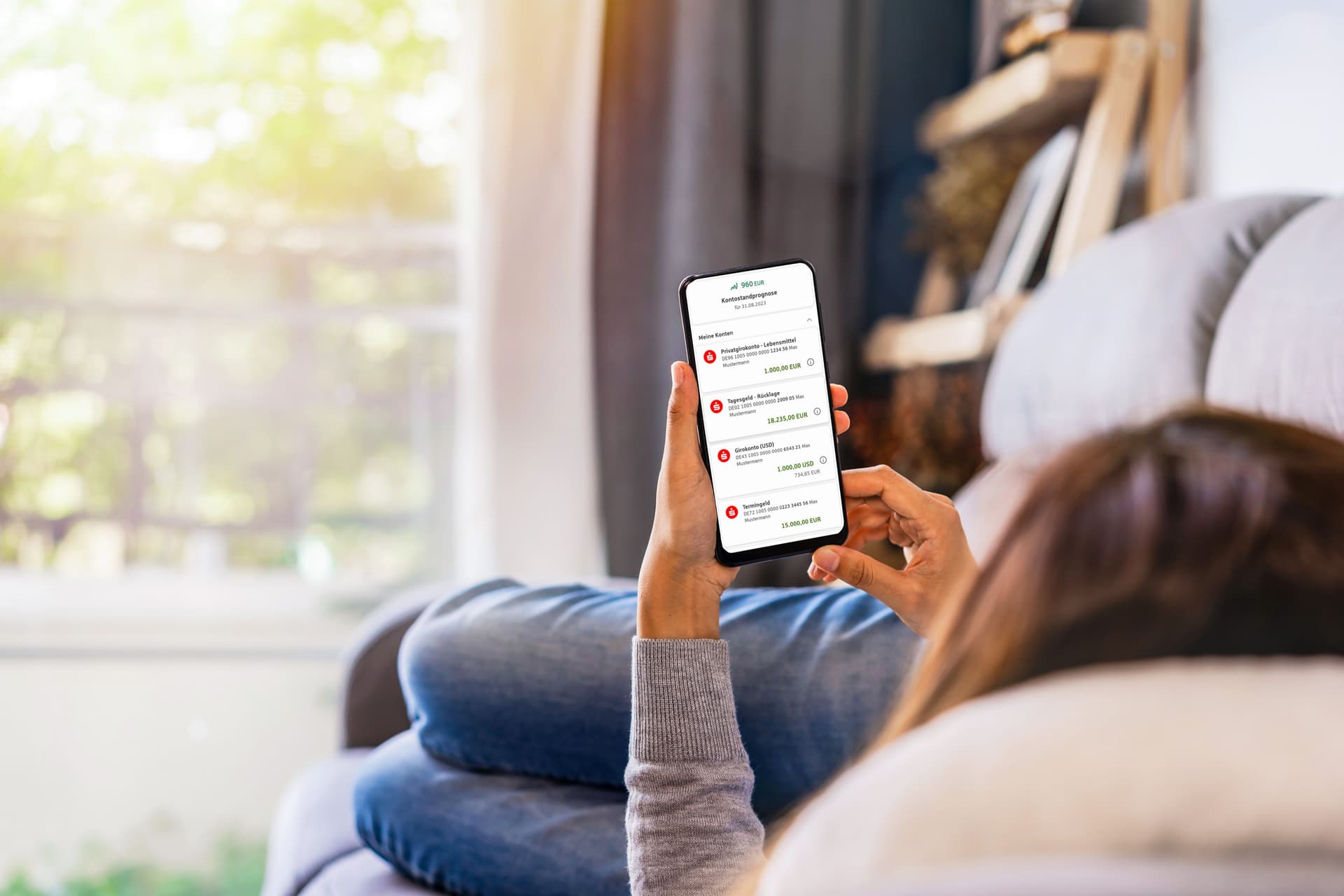 Blick über Schulter einer auf dem Sofa liegenden Person auf das Smartphone mit Banking App