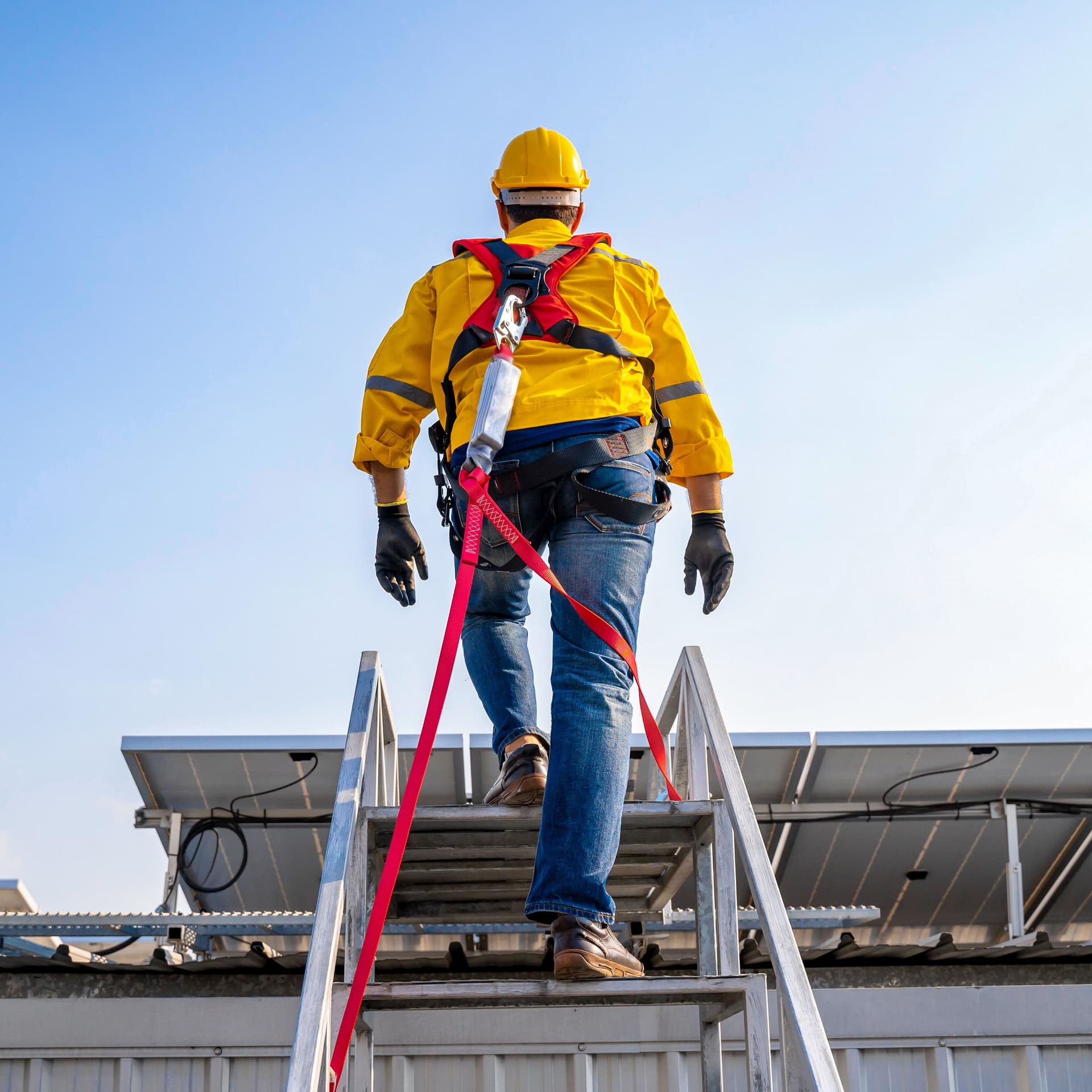 Person in gelber Arbeitsjacke und gelbem Helm steigt mit Sicherung über eine Treppe auf ein Häuserdach.