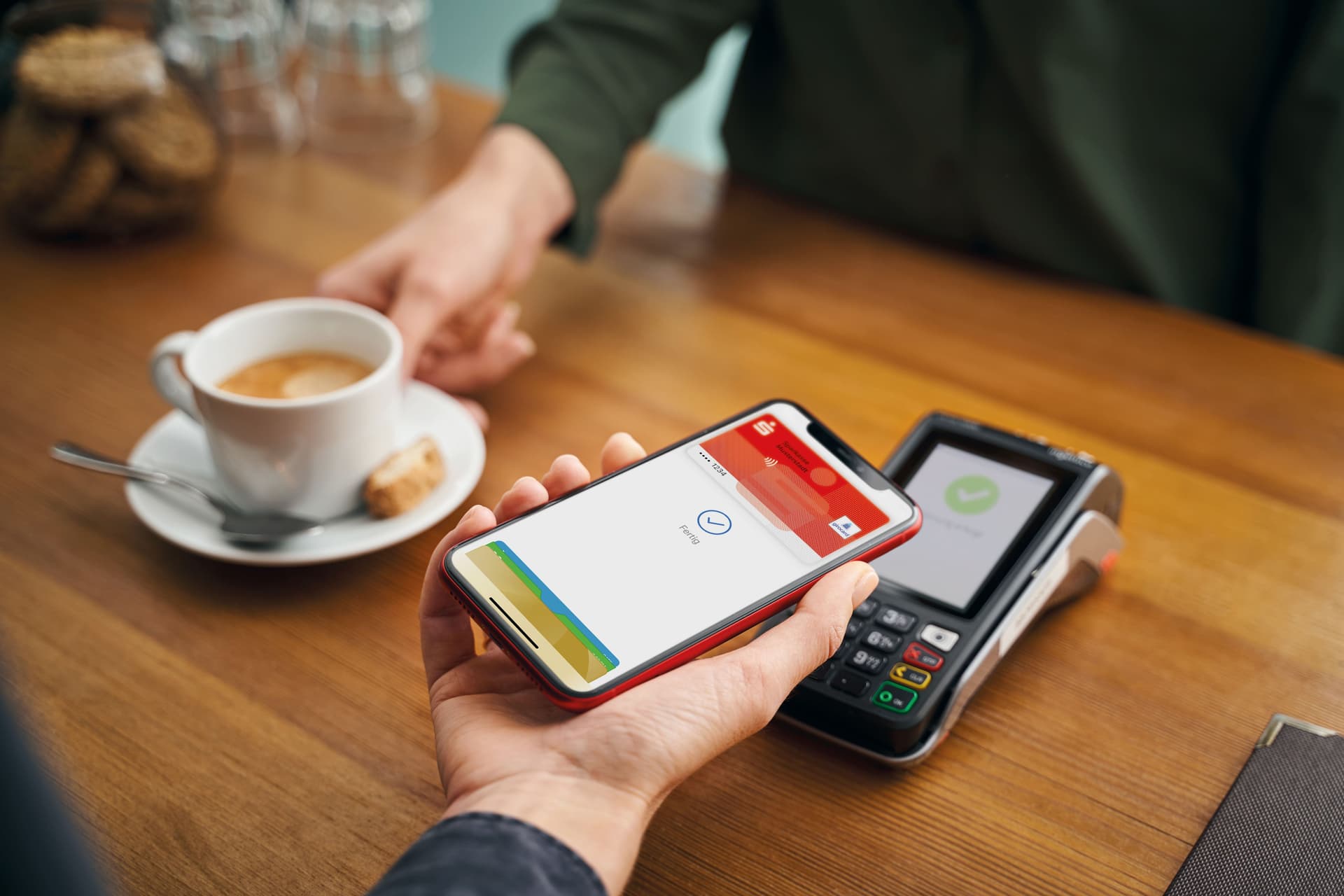 Nahaufnahe einer Hand mit Handy in einem Cafe. Auf dem Display sieht man den Bezahlvorgang für einen Kaffee über die Sparkassen-App.