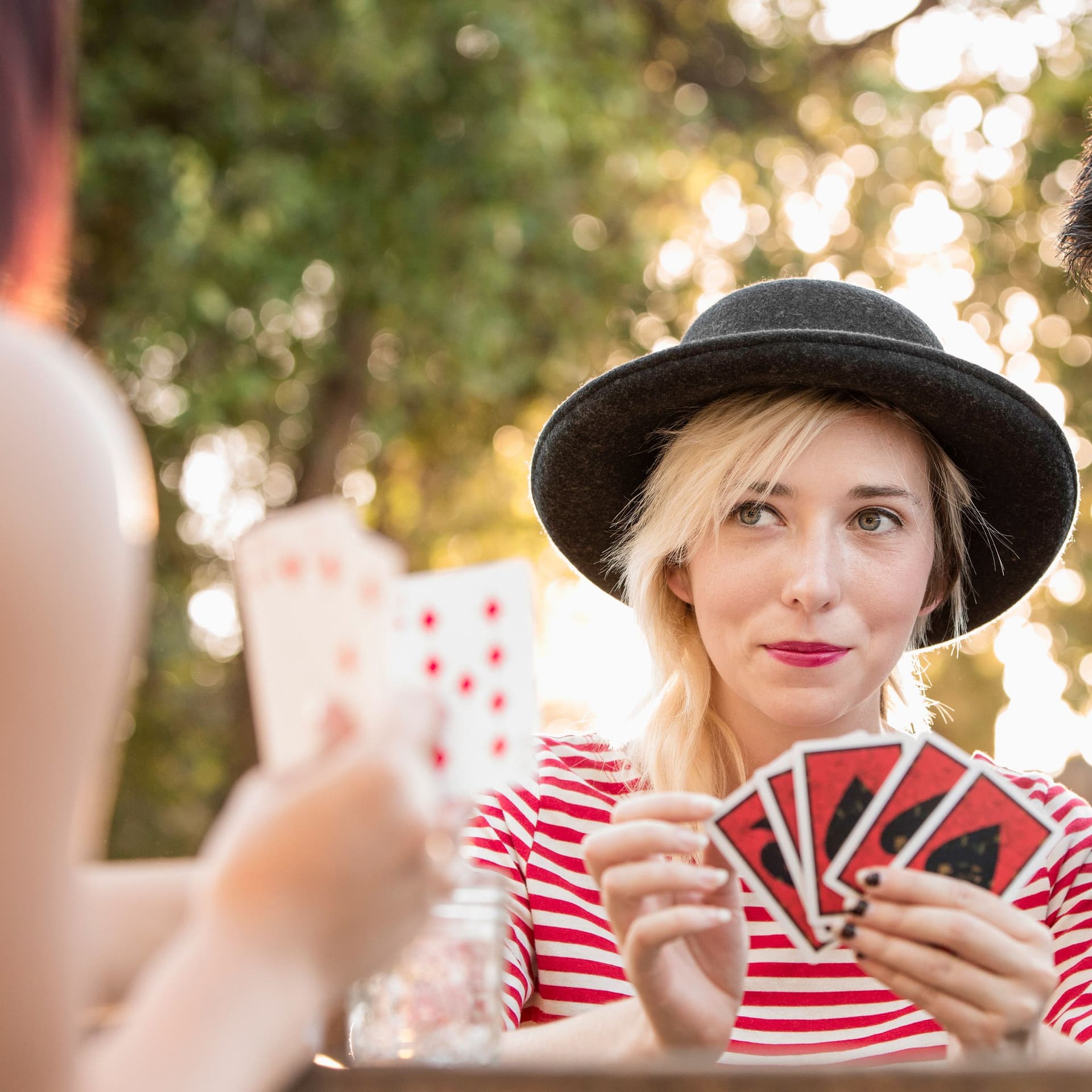 Zwei junge Frauen spielen Karten im Park. Ein Mann sitzt neben ihnen und kommentiert die Karten.