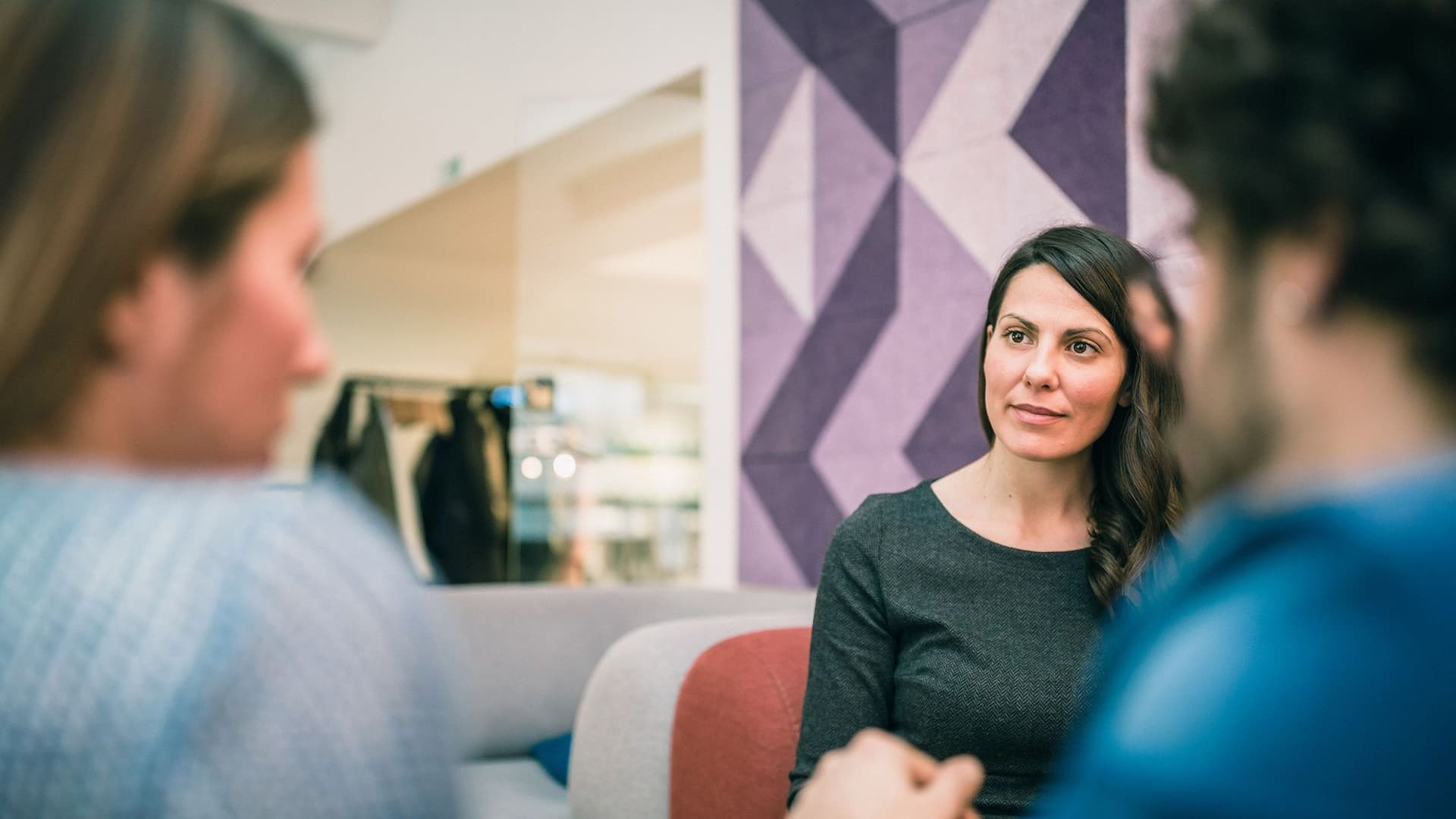 Eine Gruppe von drei Personen sitzt sich in einer Gesprächssituation gegenüber. Im Fokus steht eine junge Frau mit brauen Haaren, die mit einer vom Betrachter abgewandten Person spricht.
