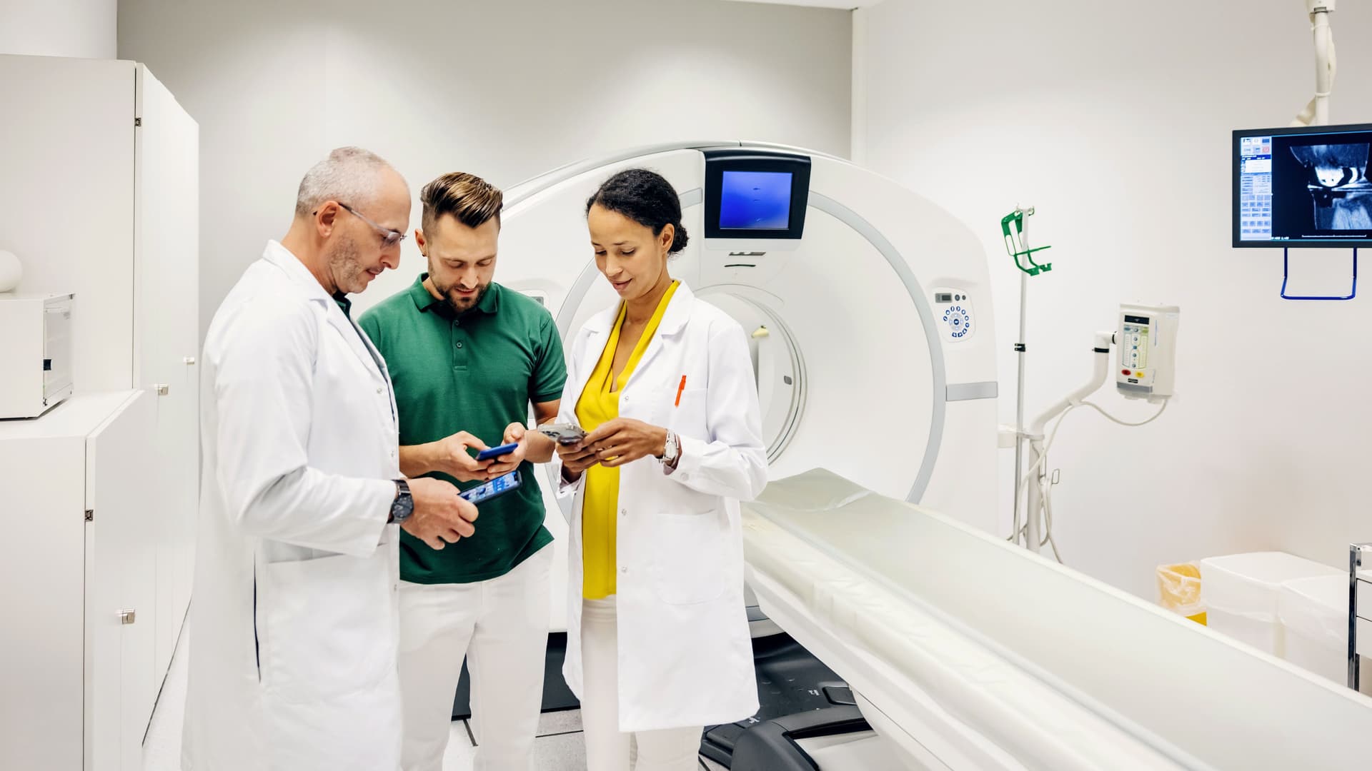 Eine Ärztin und ein Arzt besprechen einige Scandaten mit einem Mann und schauen auf ihre Smartphones, während sie in einem CT-Scanraum in einem Krankenhaus stehen.