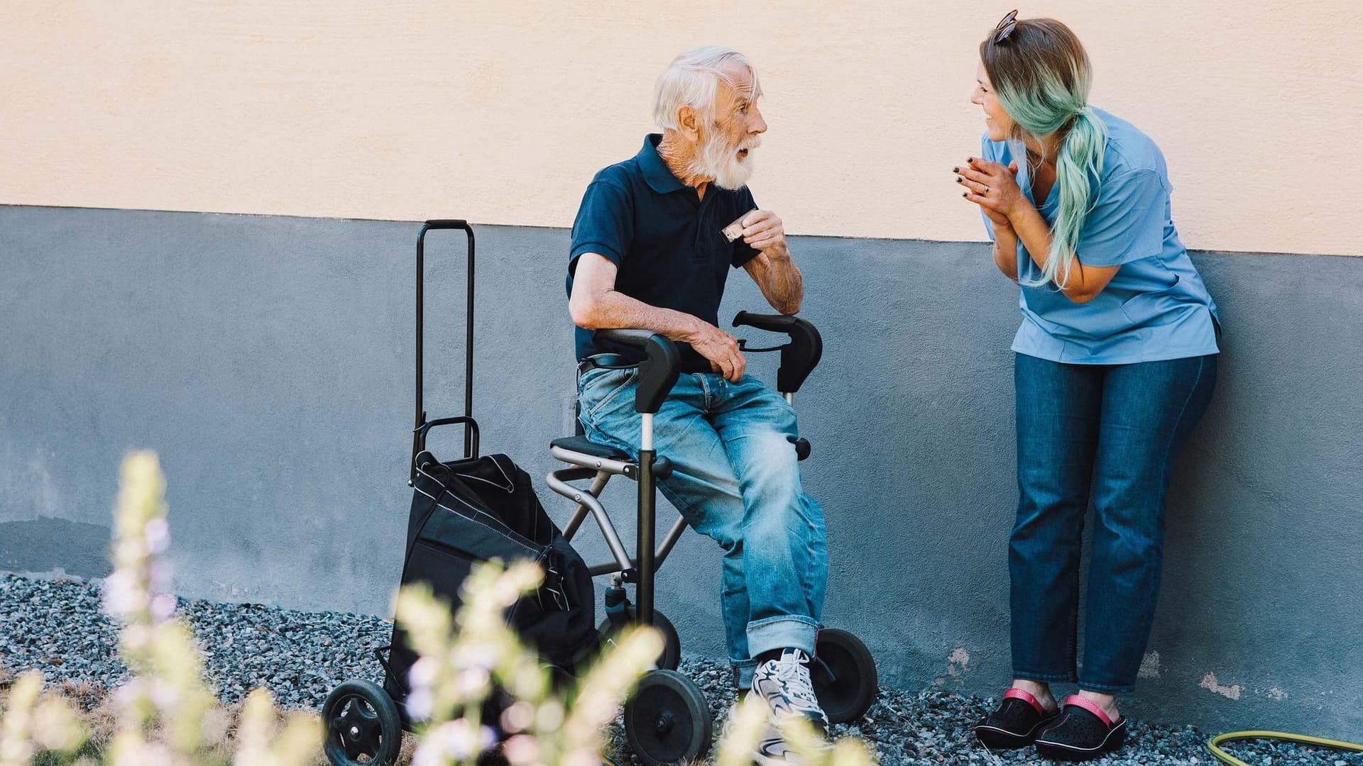 Ein älterer Mann mit weißem Haar sitzt auf einem Rollator vor einer hellen Hauswand. 