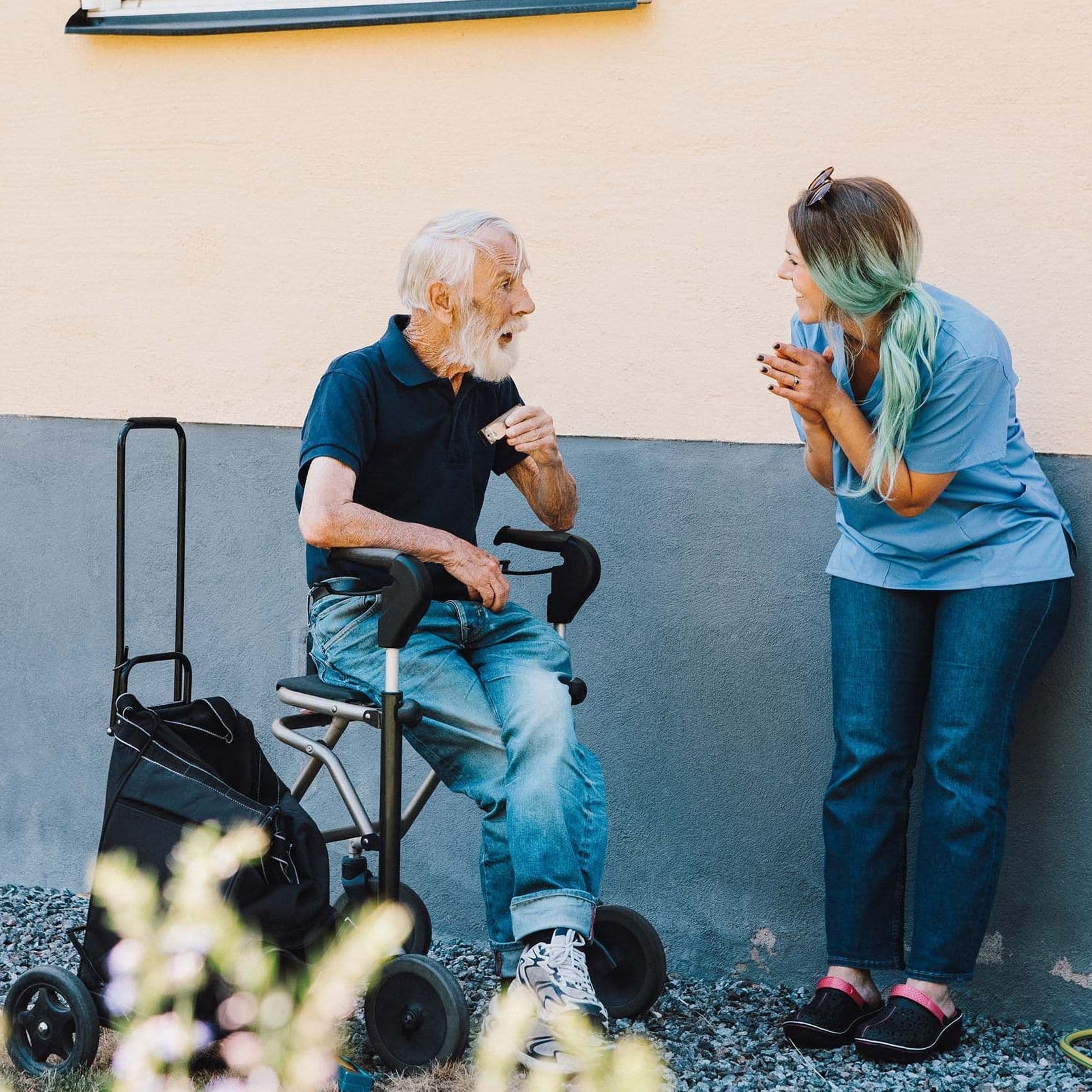 Ein älterer Mann mit weißem Haar sitzt auf einem Rollator vor einer hellen Hauswand. 
