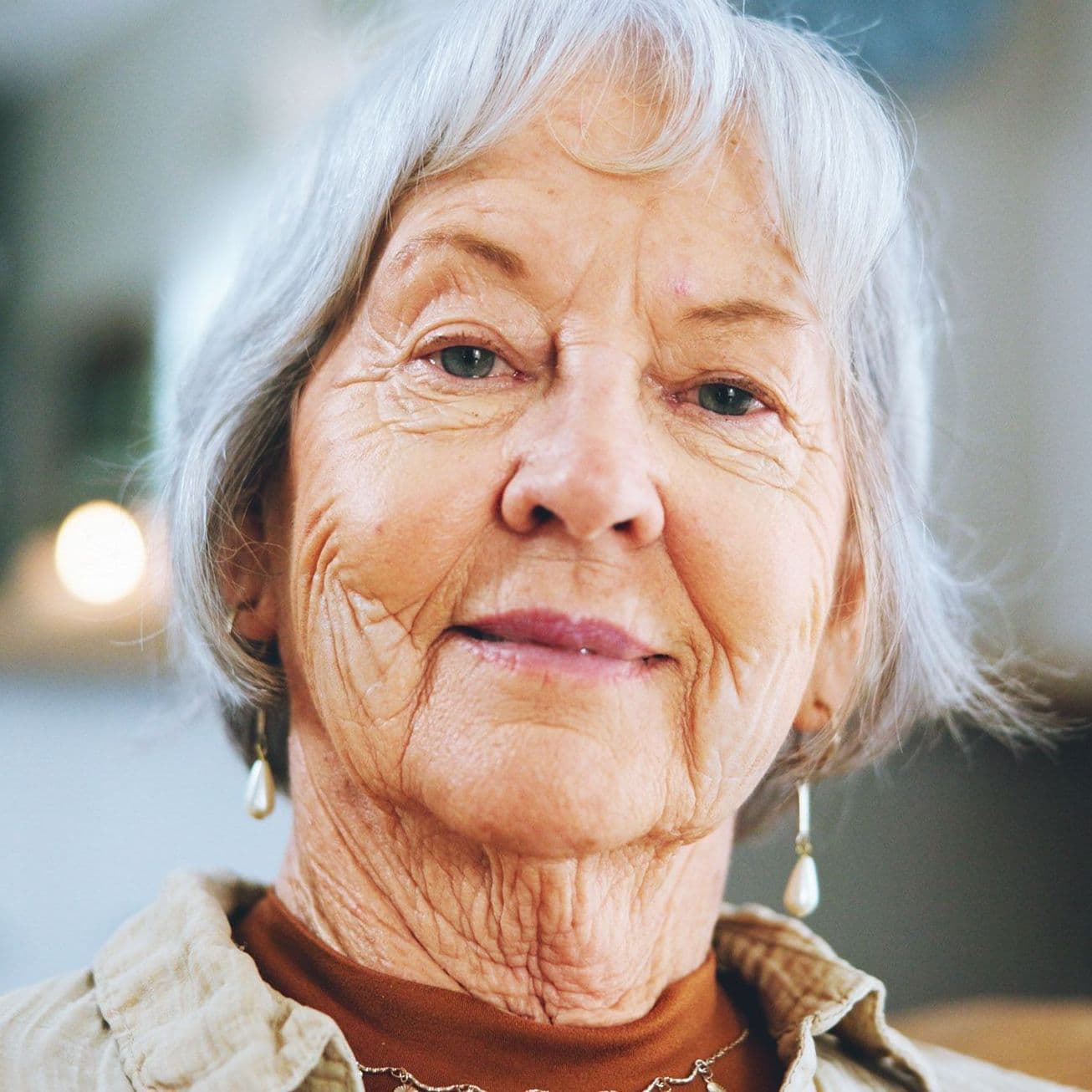 Eine Seniorin mit grauem, mittellangem Haar und freundlichem Gesichtsausdruck blickt direkt in die Kamera. Sie trägt Ohrringe und eine Kette. Der Hintergrund des Bildes ist unscharf, wodurch der Fokus ganz auf ihrem Gesicht liegt.