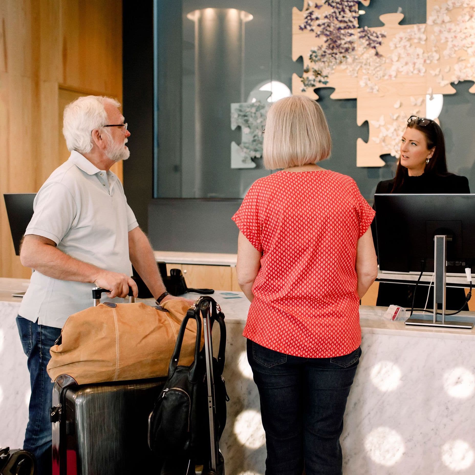 Ein älteres Paar steht mit seinem Urlaubsgepäck in einer Hotellobby und führt ein Gespräch mit der Rezeptionistin.