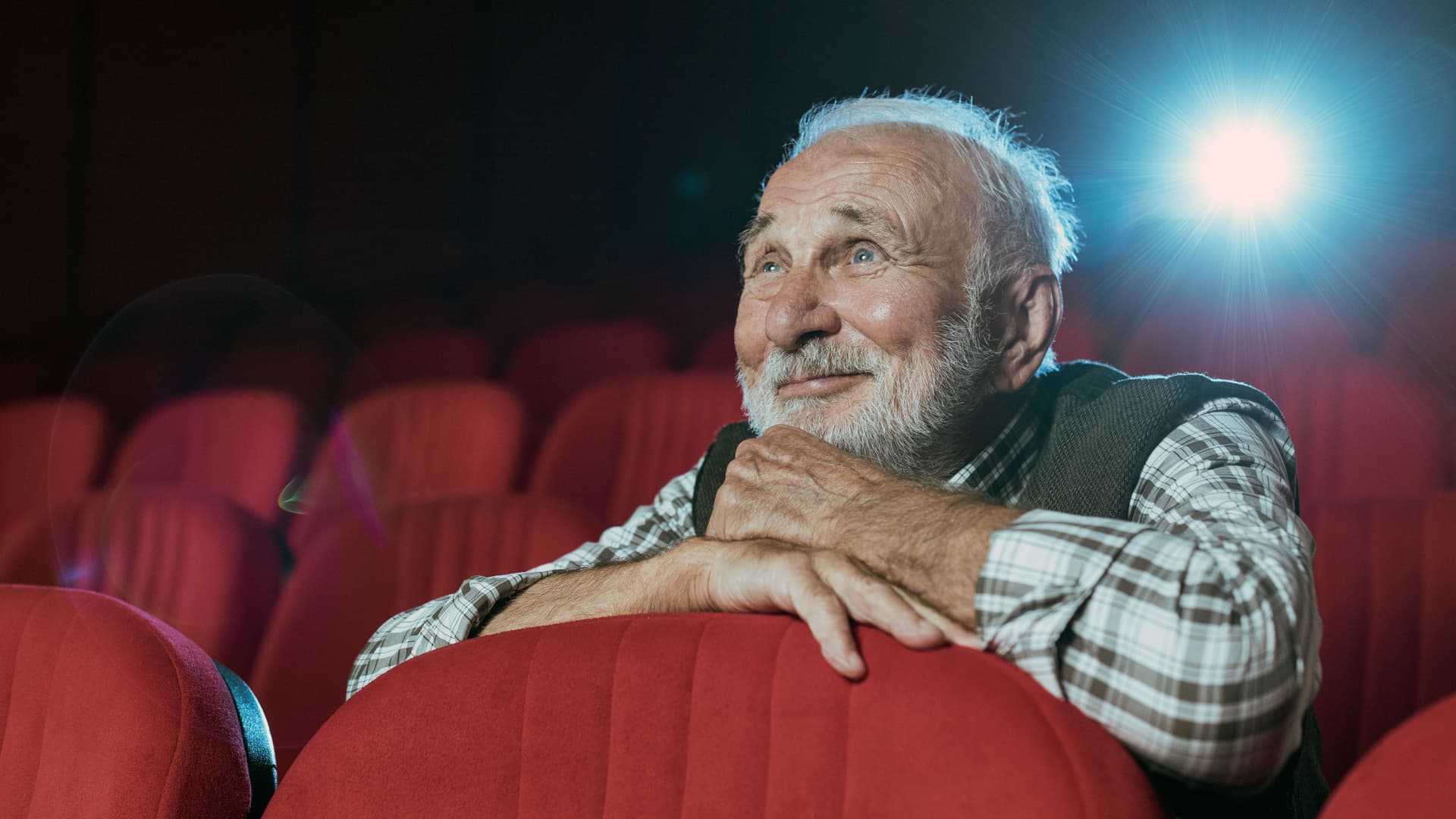 Ein Senior mit grauen Haaren und Bart sitzt in einem Kino und schaut lächelnd einen Film.