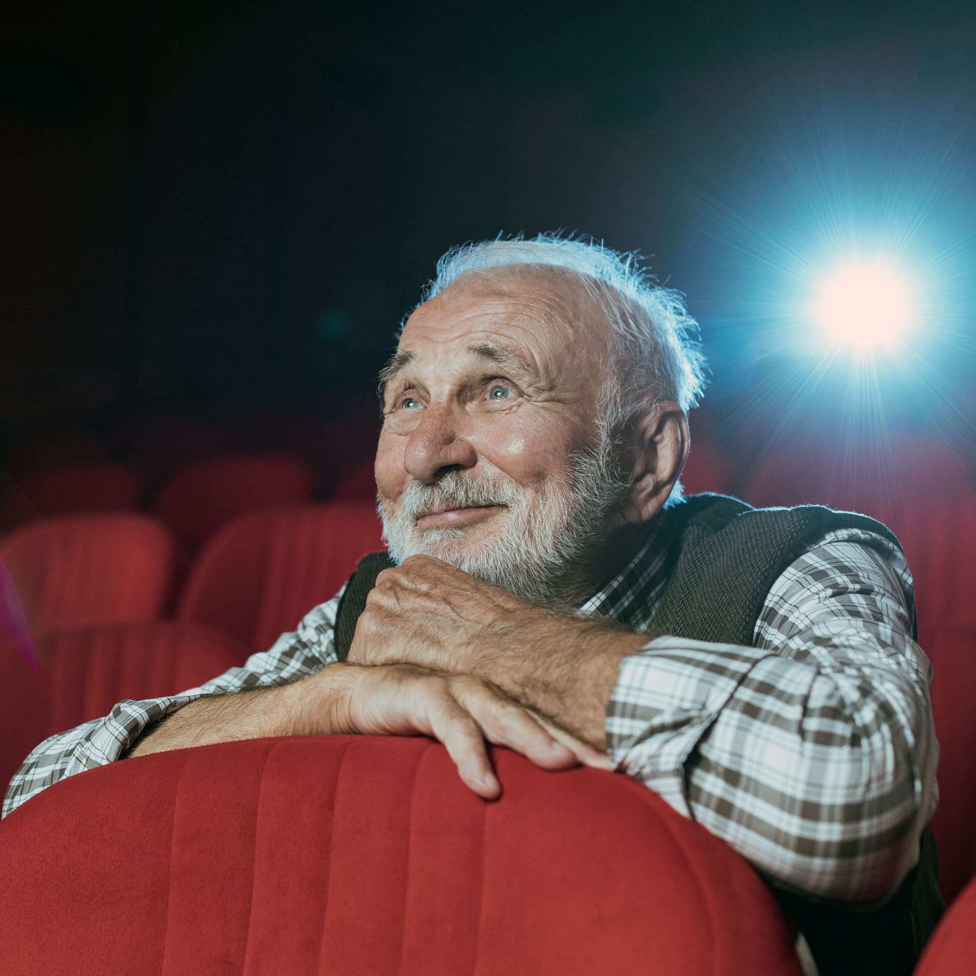 Ein Senior mit grauen Haaren und Bart sitzt in einem Kino und schaut lächelnd einen Film.