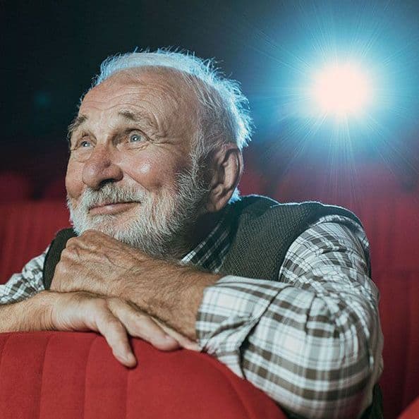 Ein Senior mit grauen Haaren und Bart sitzt in einem Kino und schaut lächelnd einen Film.