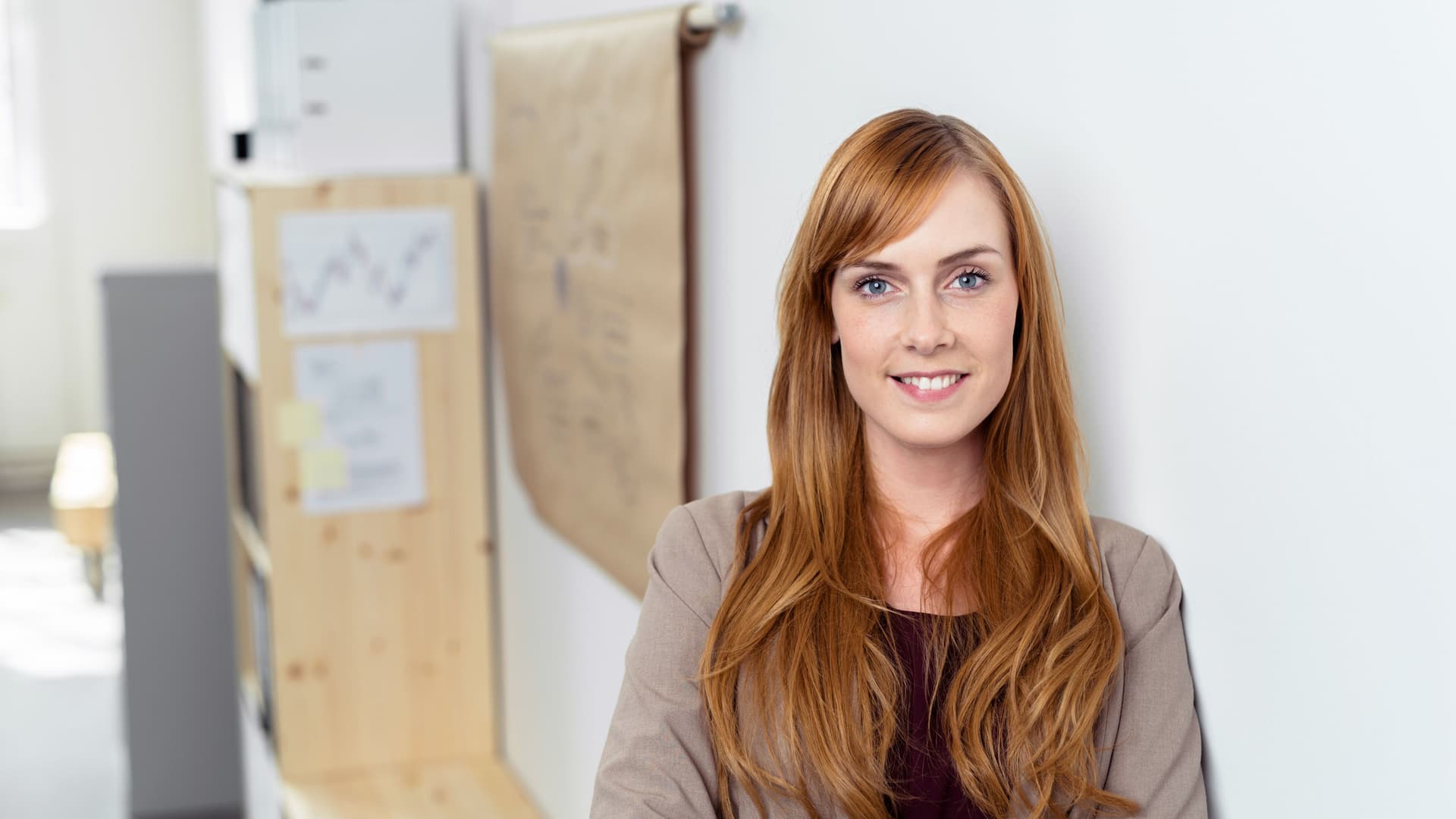 Junge Frau steht an eine Wand gelehnt in einem Büro und lächelt in die Kamera.