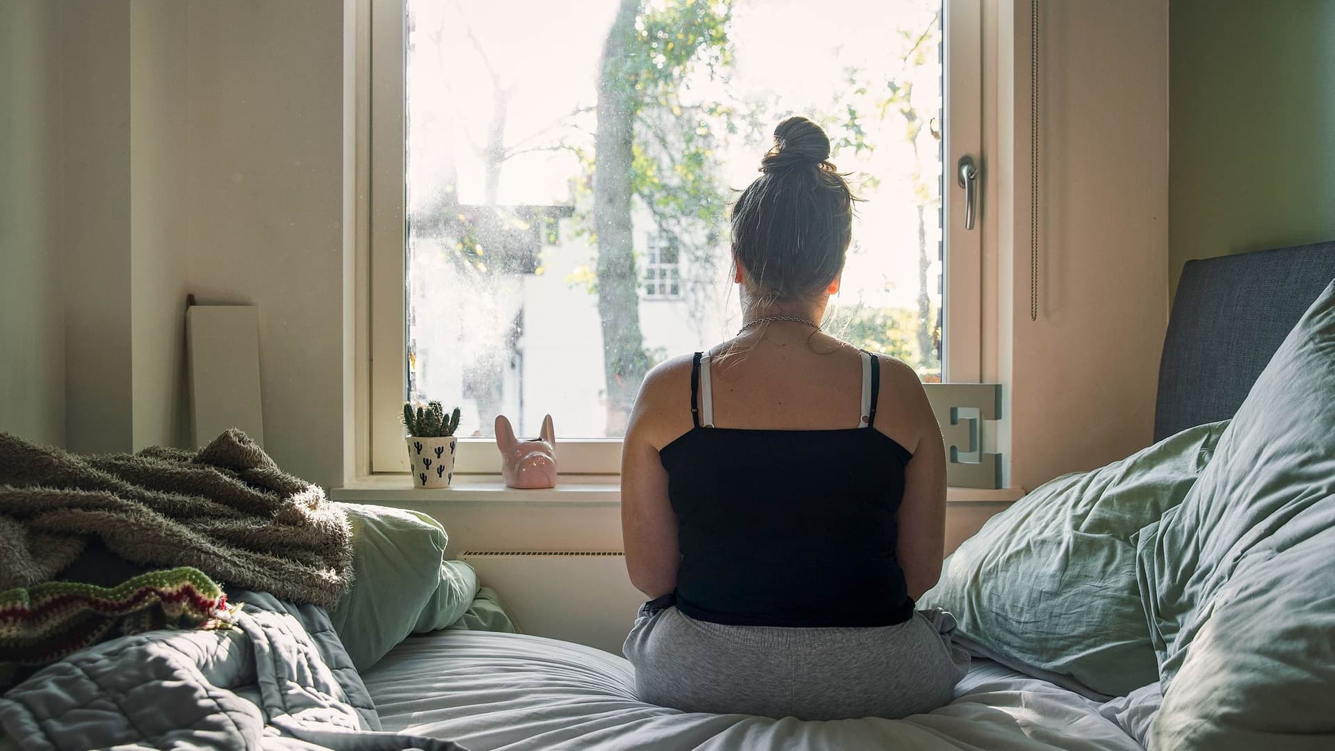 Eine Frau sitzt mit dem Rücken zur Kamera auf einem Bett und blickt aus dem Fenster. Das Tageslicht fällt in den Raum und schafft eine ruhige, nachdenkliche Atmosphäre.