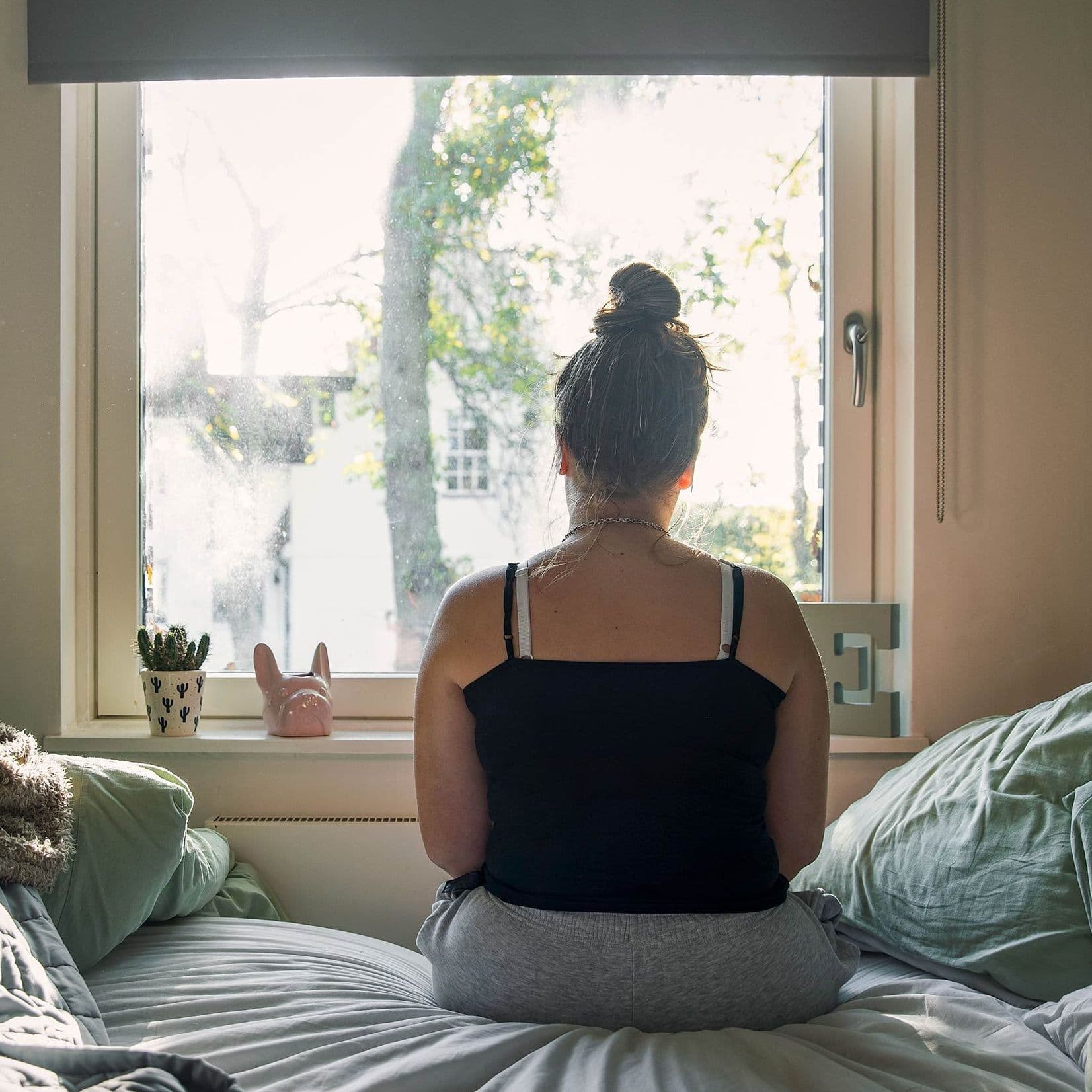 Eine Frau sitzt mit dem Rücken zur Kamera auf einem Bett und blickt aus dem Fenster. Das Tageslicht fällt in den Raum und schafft eine ruhige, nachdenkliche Atmosphäre.
